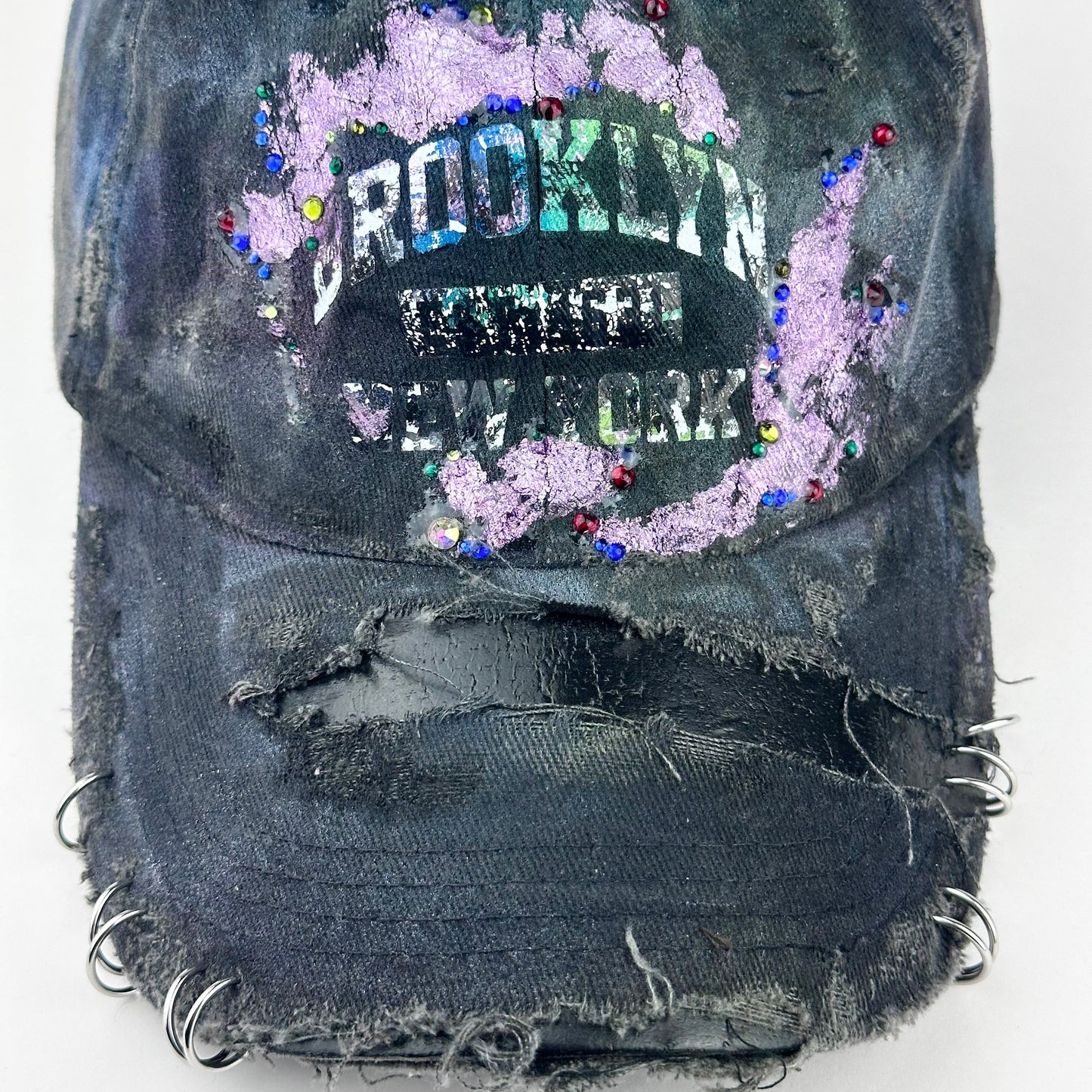 Close up of ASTROBOI* distressed black Brooklyn hat with metallic purple paint, multicolored rhinestones, green airbrush shading, and white front lettering.
