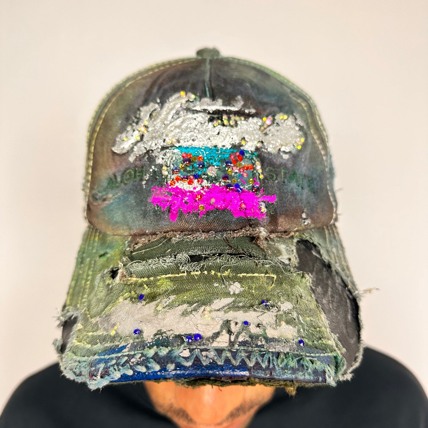 On body front shot of Brown-green ASTROBOI* hat featuring a metallic Hawaii logo with multicolored rhinestones, distressed visor, and resin-coated safety pin detail.