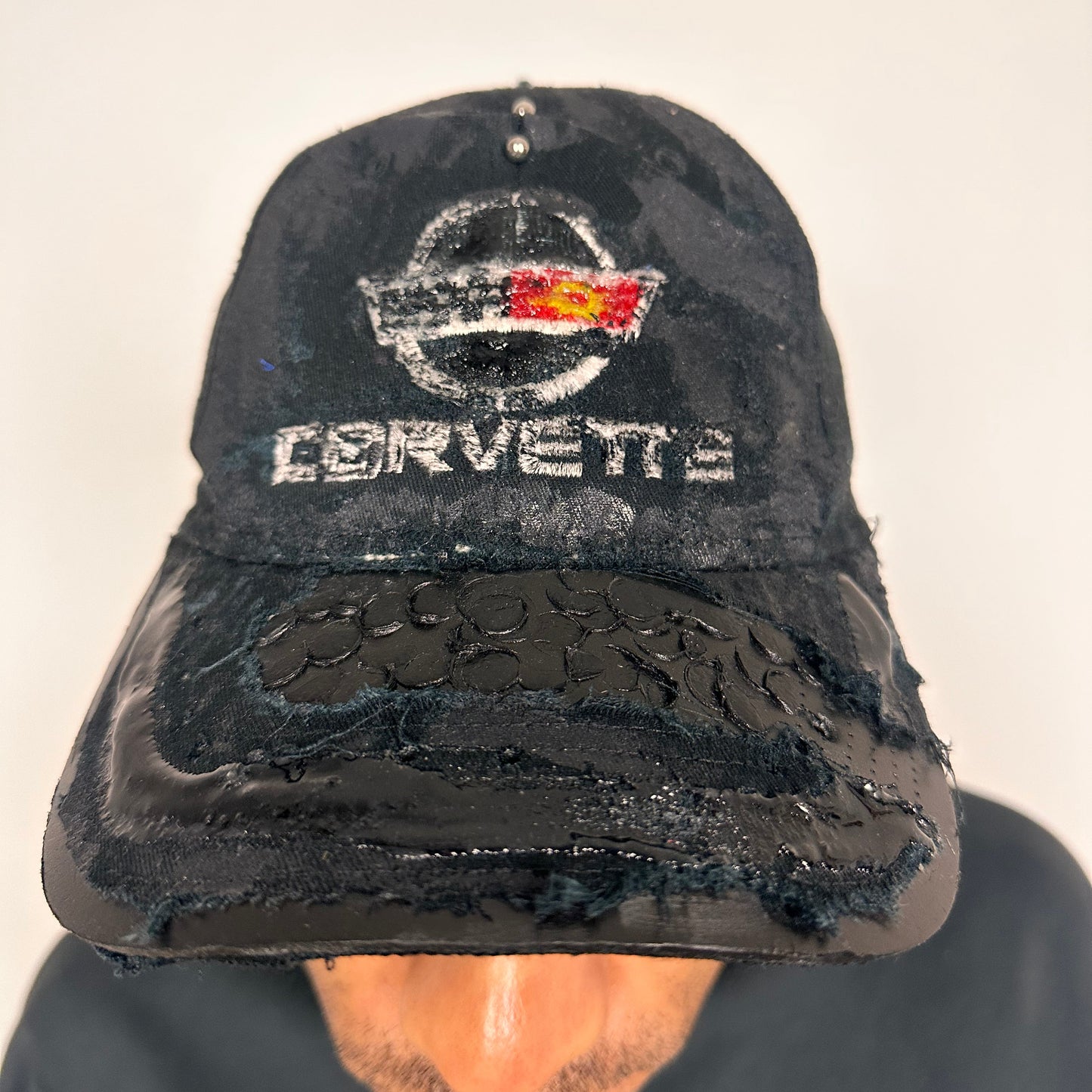 Front close up of “Black distressed ASTROBOI* x Corvette hat with resin-coated logo, stainless steel piercings, and deconstructed visor with circular peep holes.”