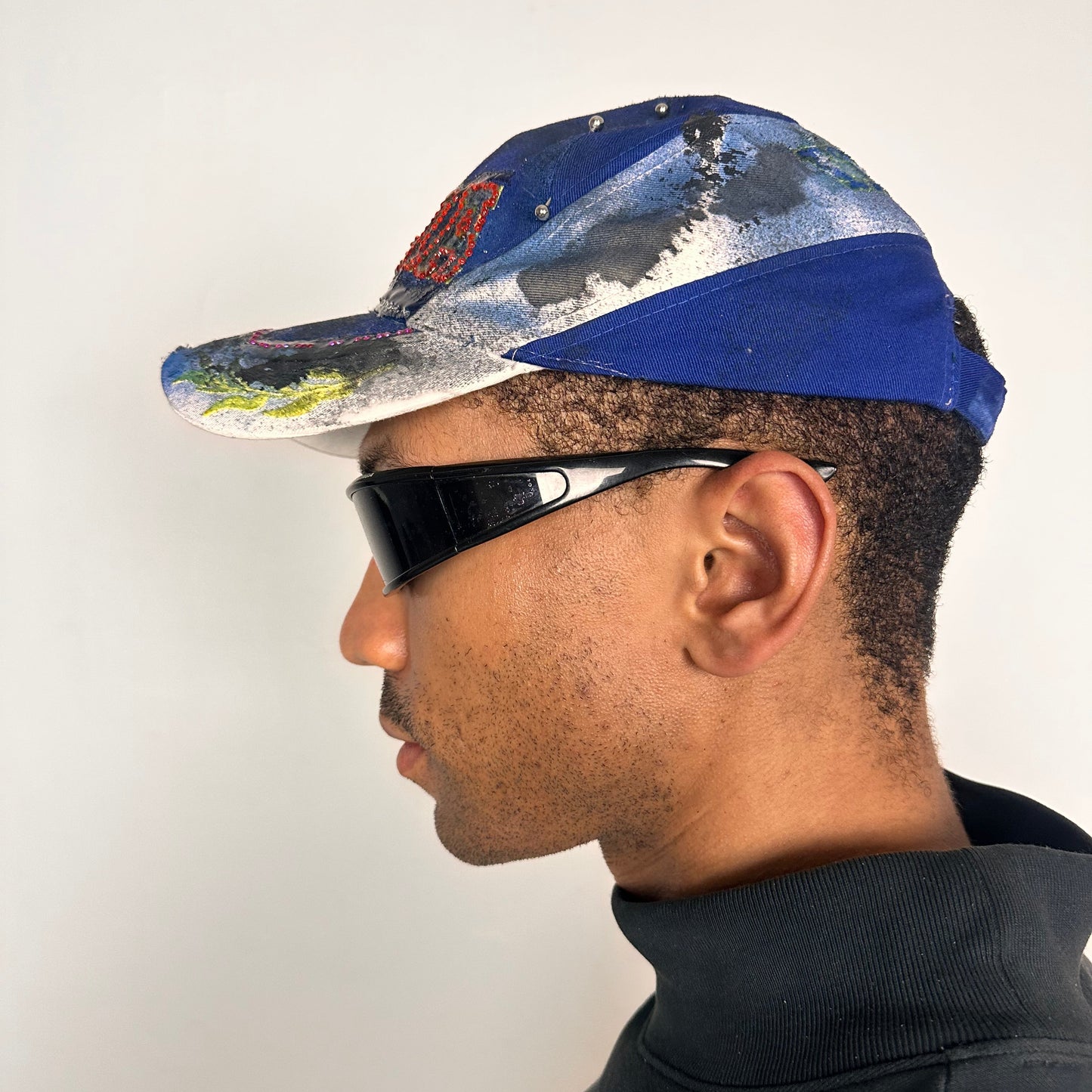 Left of Upcycled blue and white ASTROBOI* ‘ITALIA’ hat with rhinestones, resin, custom paint, and stainless steel piercings, handcrafted in NYC.