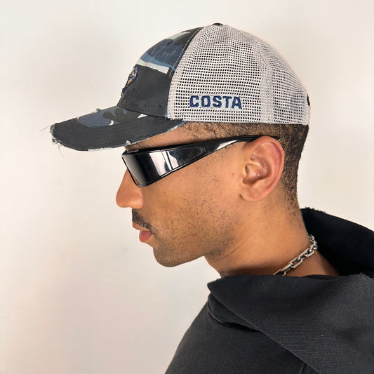 Left side of ASTROBOI* Costa blue and white camo rhinestone trucker hat with black waxed tattoo print, mesh back panels, adjustable strap, custom NYC piece