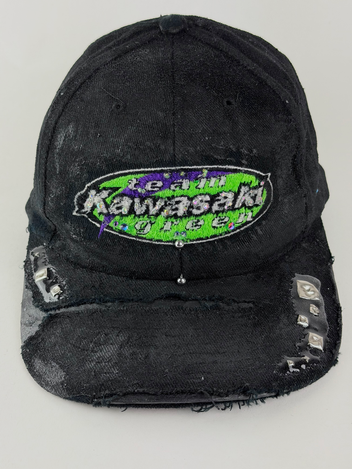 ASTROBOI* x Kawasaki 'Faust' black green and purple rhinestone studded cap with rhinestone-embellished Kawasaki logo, stainless steel piercing, and deconstructed visor, front view close up.