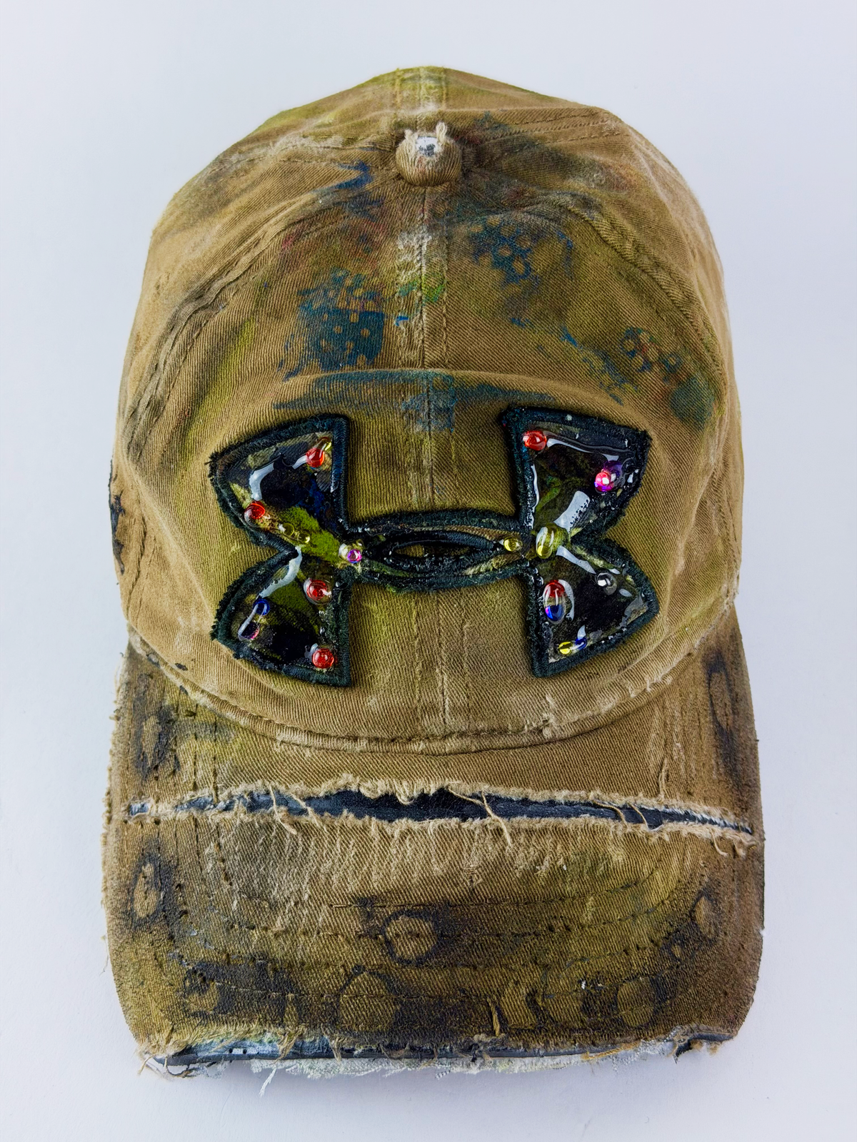 A distressed olive green baseball cap featuring a large, embellished Under Armour logo patch on the front, set against a plain white background.