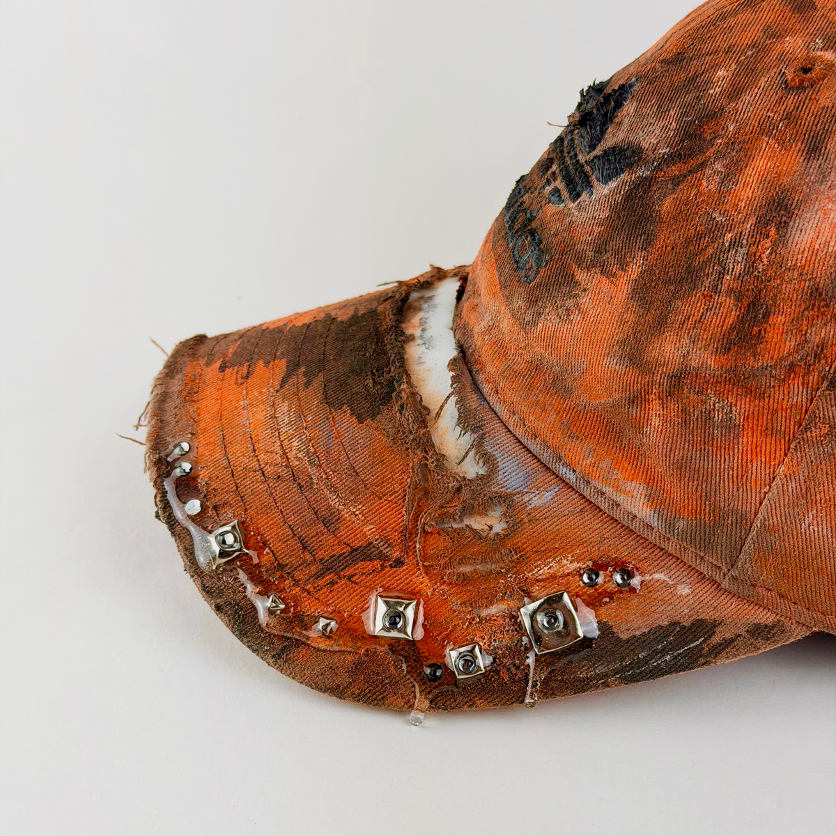 A close-up of a distressed orange baseball cap with heavy rust and black staining, featuring a tattered brim adorned with small silver metal studs and clear resin details, set against a plain light gray background.