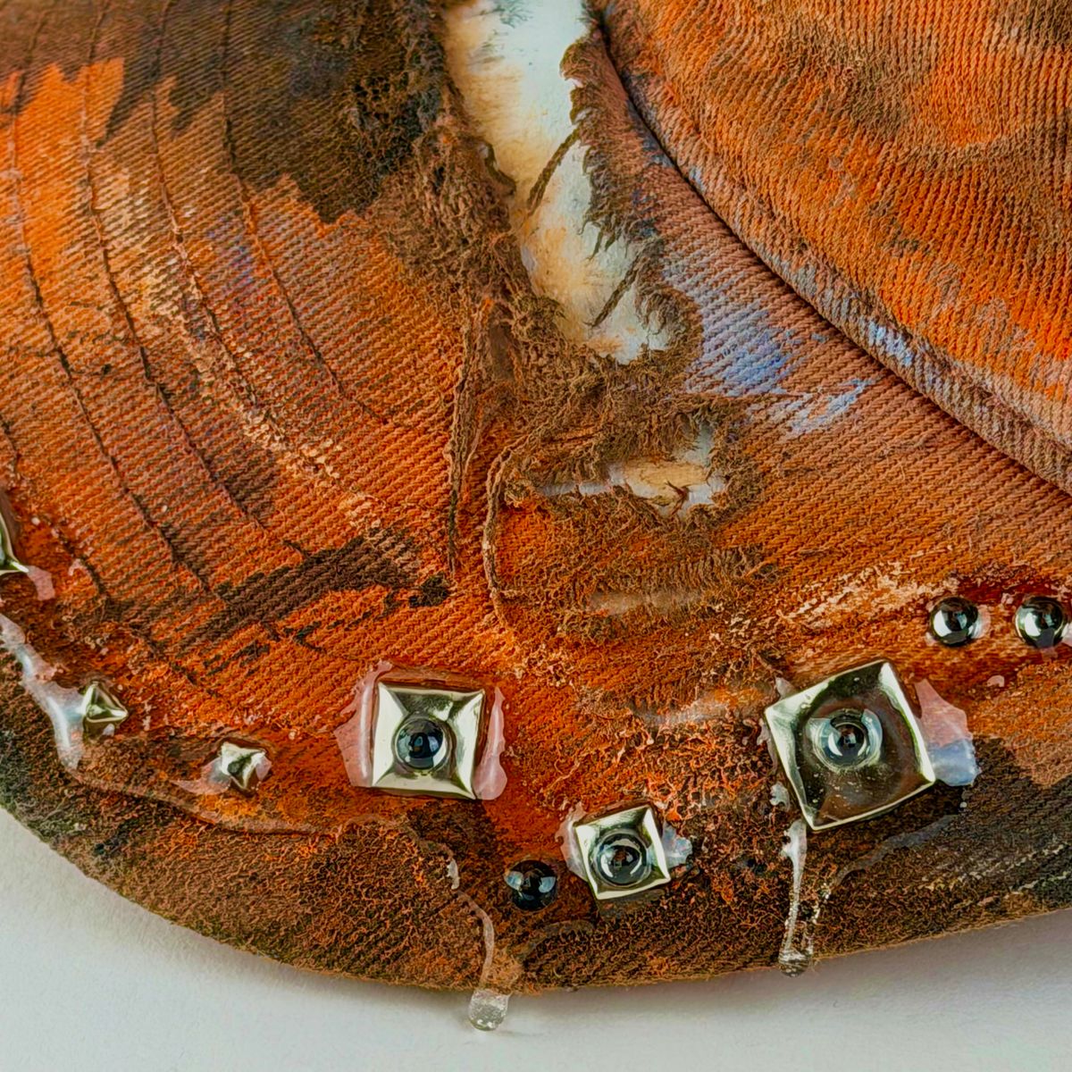 Close-up of a distressed orange fabric surface with frayed texture, silver studs, rhinestones, and clear resin detailing.