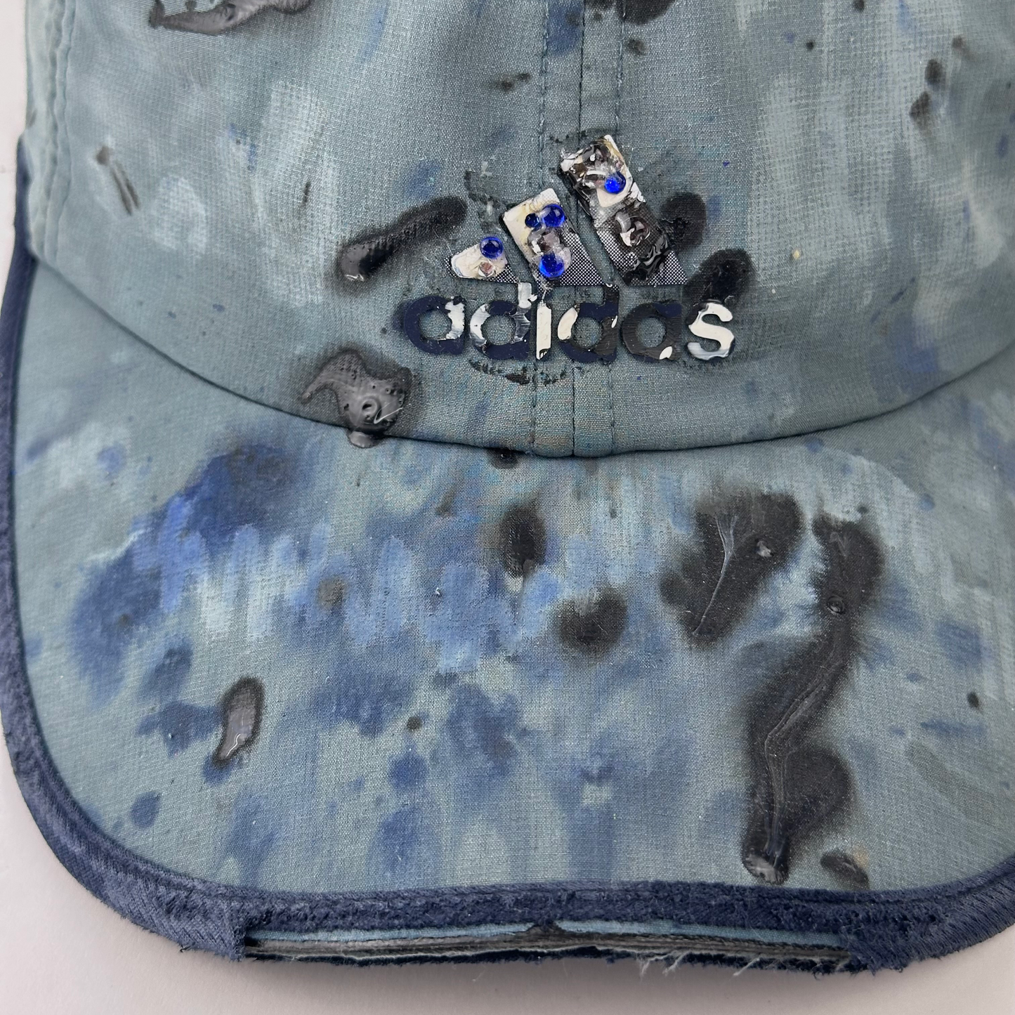 Close-up of ASTROBOI* x Adidas 'Haaland' showing rhinestone-embellished logo detail and faded black, white, and blue raised paint treatment on moisture-wicking fabric.