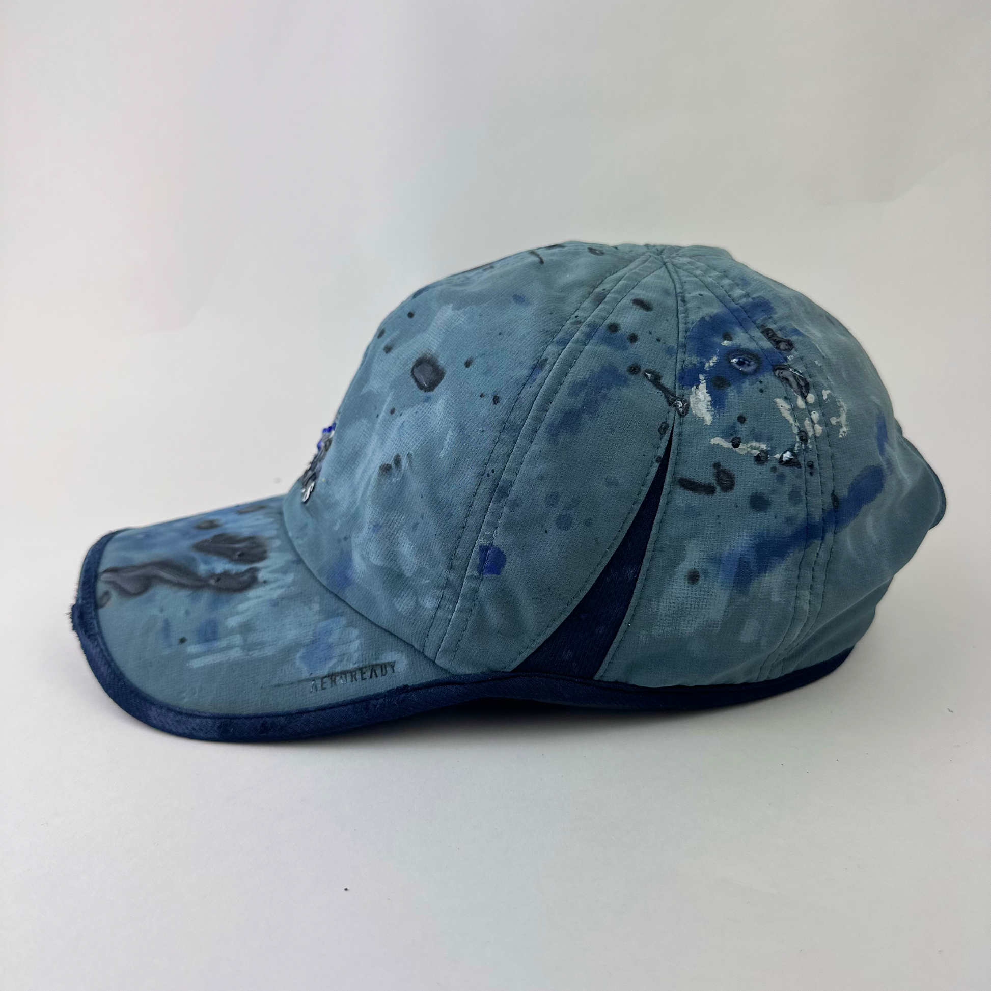 Left side detail of ASTROBOI* x Adidas 'Haaland' showing raised faded paint treatment and blue visor trim detail on blue and black cap.