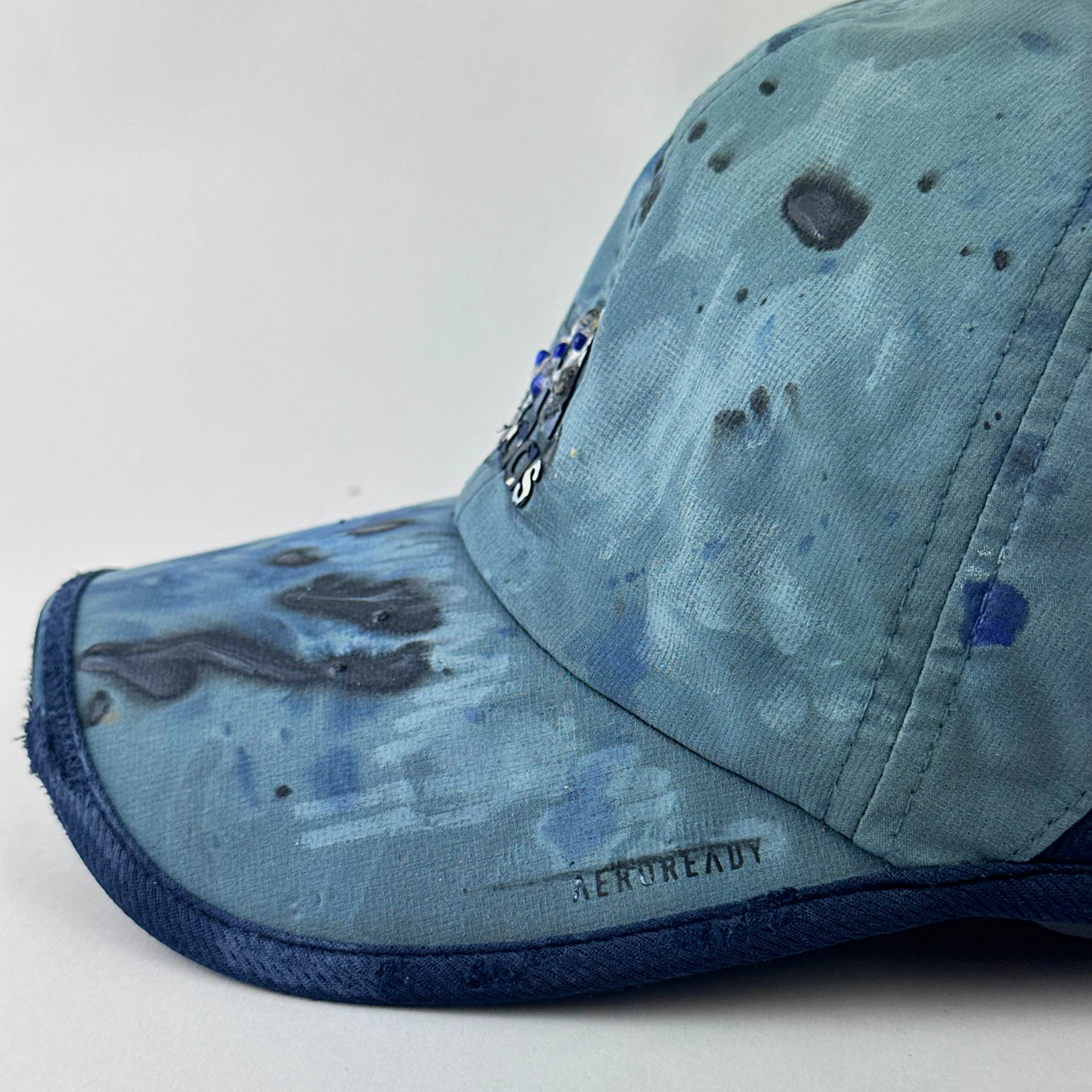 Aeroready detail of ASTROBOI* x Adidas 'Haaland' showing raised faded paint treatment and blue visor trim detail on blue and black cap.