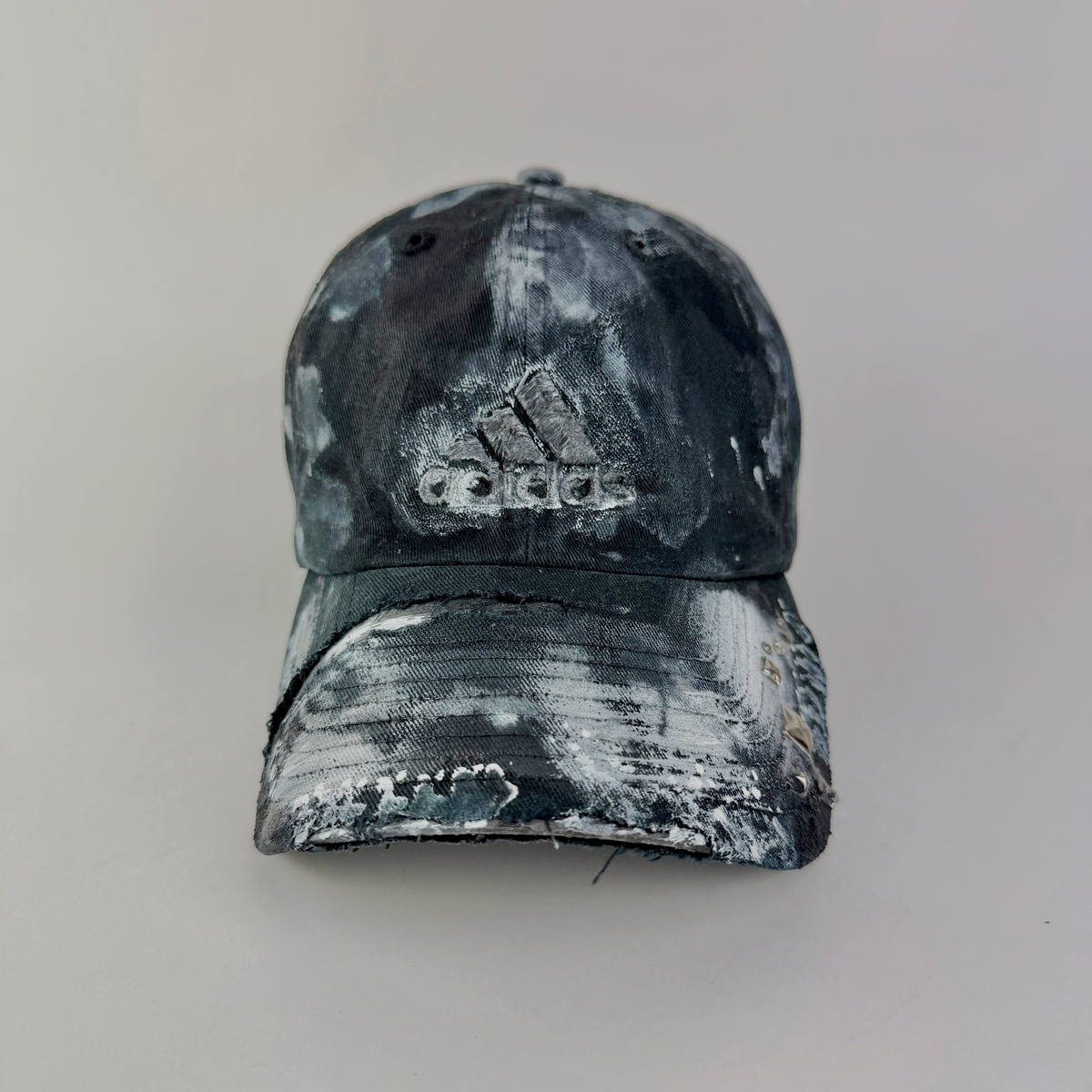 A distressed Adidas baseball cap with a black and white paint-splatter or acid-wash pattern, shown against a plain light gray background.
