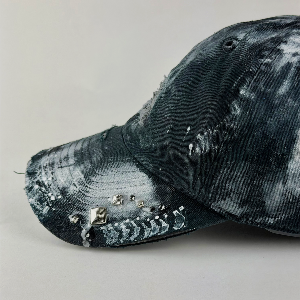 A close-up side view of a distressed baseball cap in black and white tie-dye, decorated with rhinestones and studs on the brim, set against a plain light gray background.