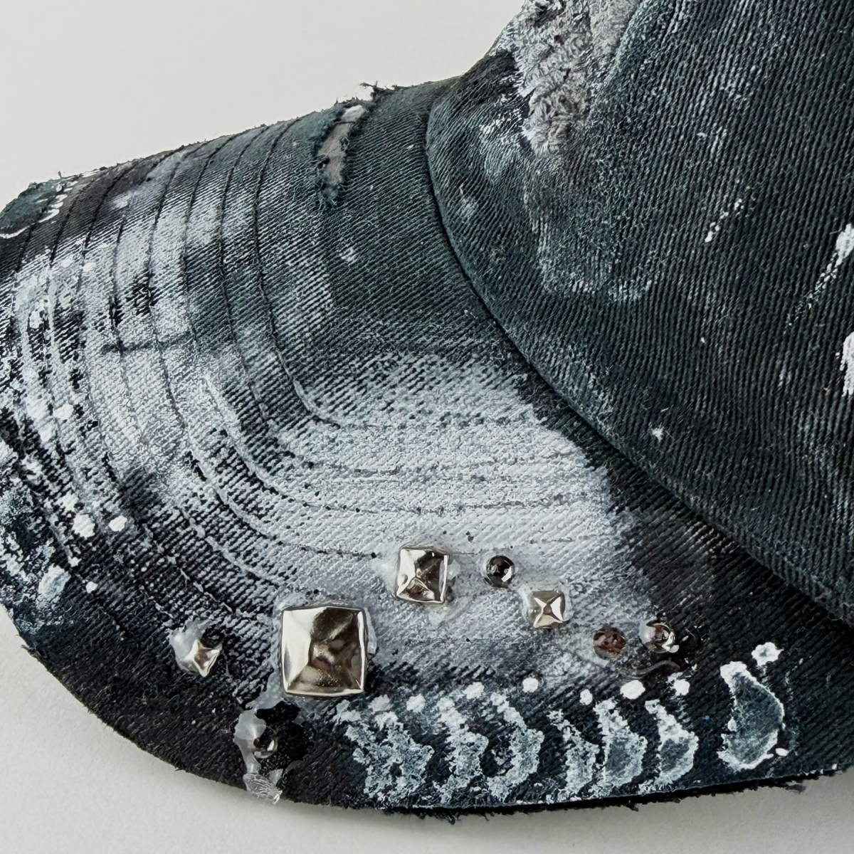 Close-up of a distressed baseball cap in black and white paint splatter fabric, decorated with small metallic studs and rhinestones on the brim.