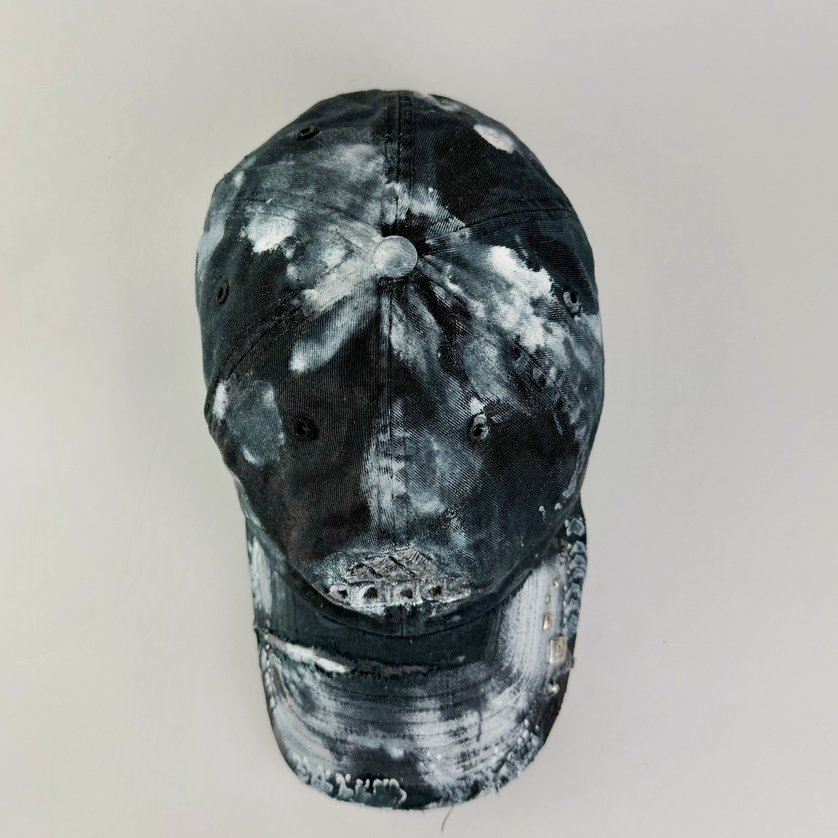 A distressed baseball cap featuring a black and white tie-dye or bleach-splattered pattern, shown against a plain light gray background.