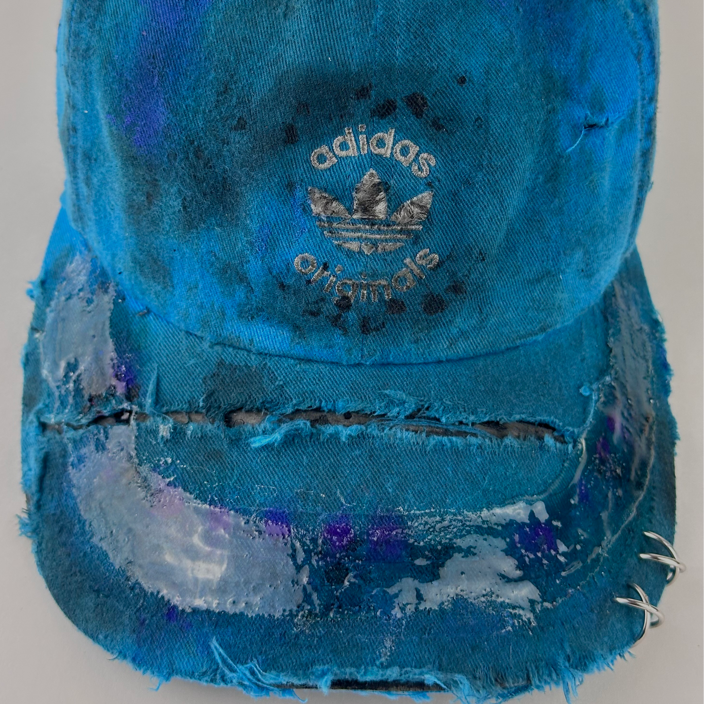 Close-up of ASTROBOI* x Adidas 'Nebula' showing white Adidas logo, stainless steel piercing detail, and resin 3D finish along the visor on distressed blue fabric.