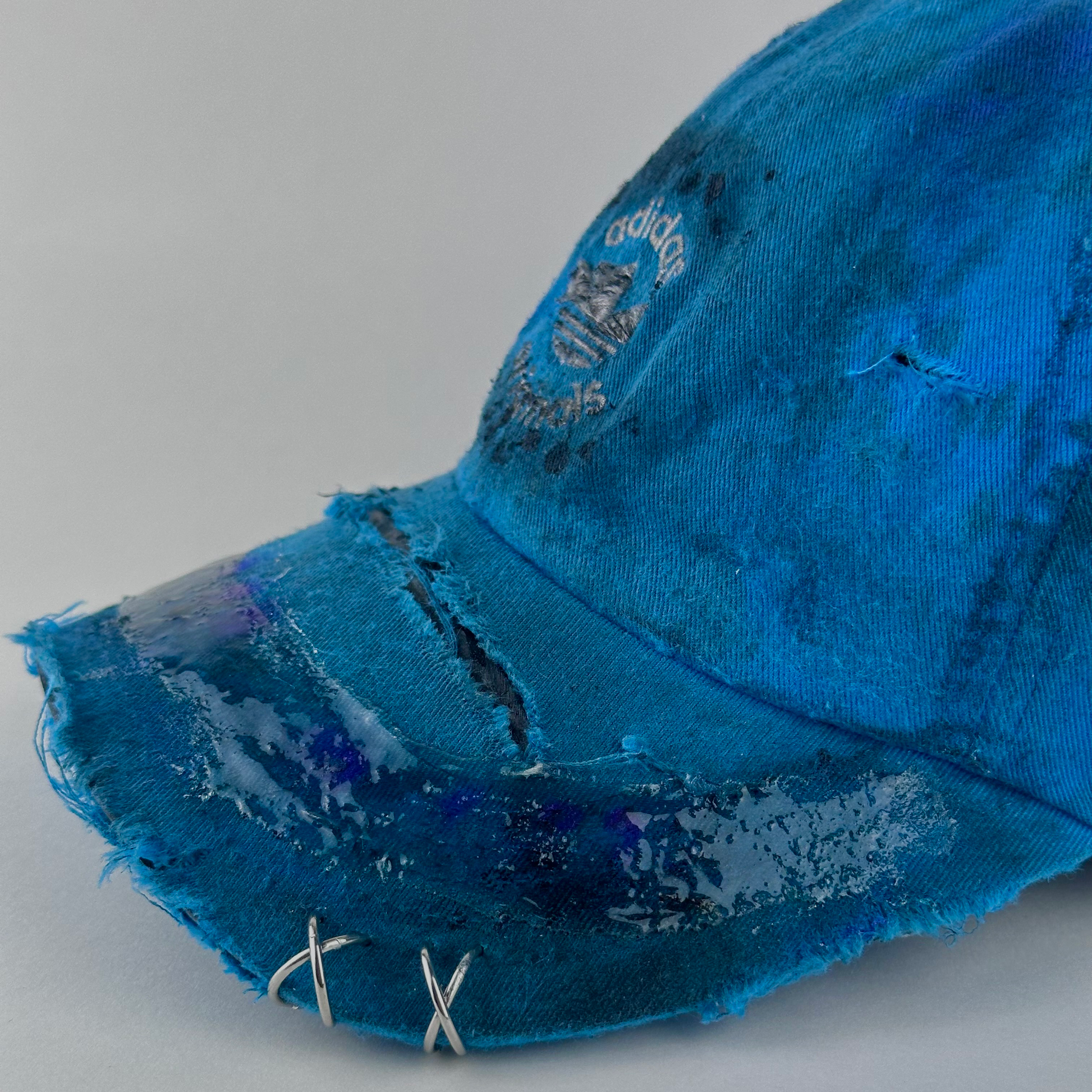 Left side piercing detail of ASTROBOI* x Adidas 'Nebula' showing distressing, custom paint treatment, and resin visor detail on blue Adidas hat.