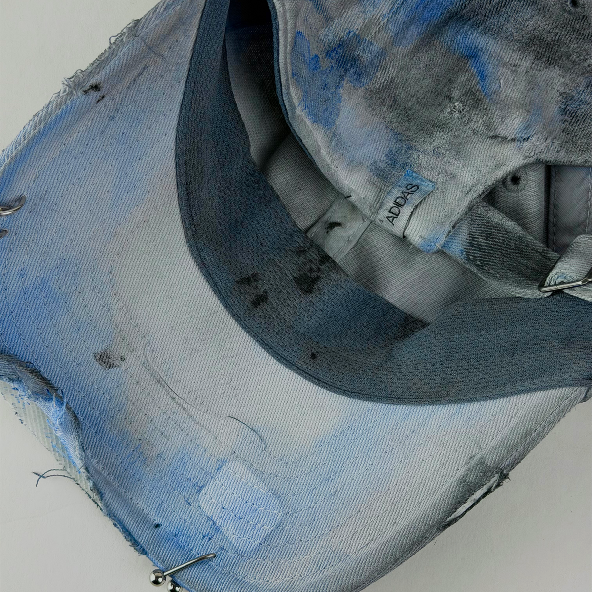 A close-up of a distressed denim baseball cap with heavy blue and gray bleach washes, frayed edges, and metal piercings attached to the brim and crown. 