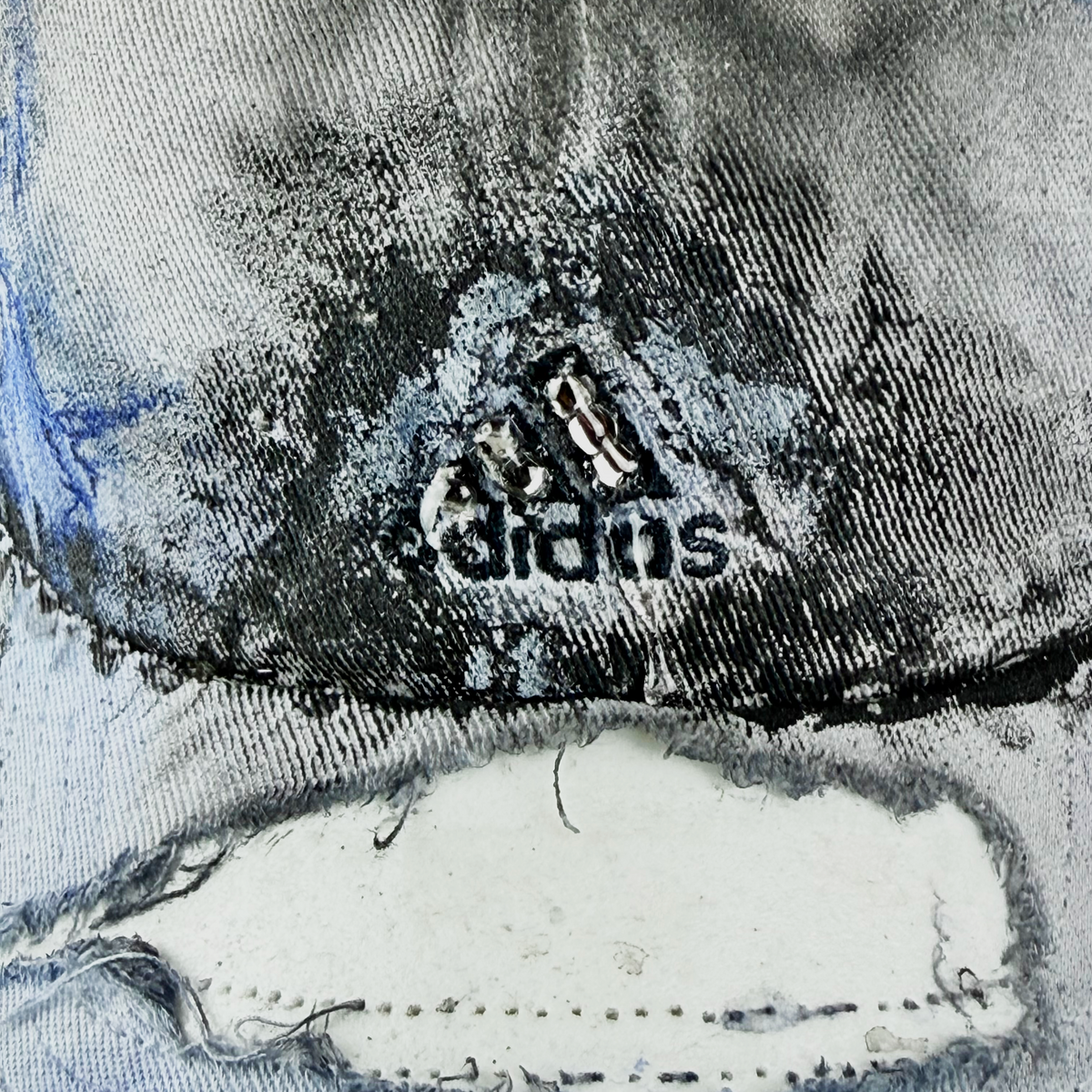 Close-up of a distressed denim product with heavy fading, a large ripped area, and a visible Adidas logo patch near the center.