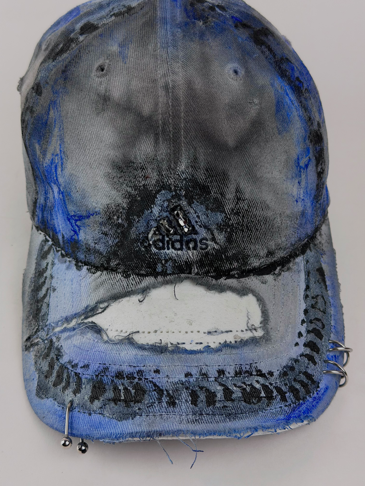 A distressed Adidas baseball cap with heavy blue and black paint splatter and chipping, featuring a large white patch on the brim and metal eyelets on the sides.