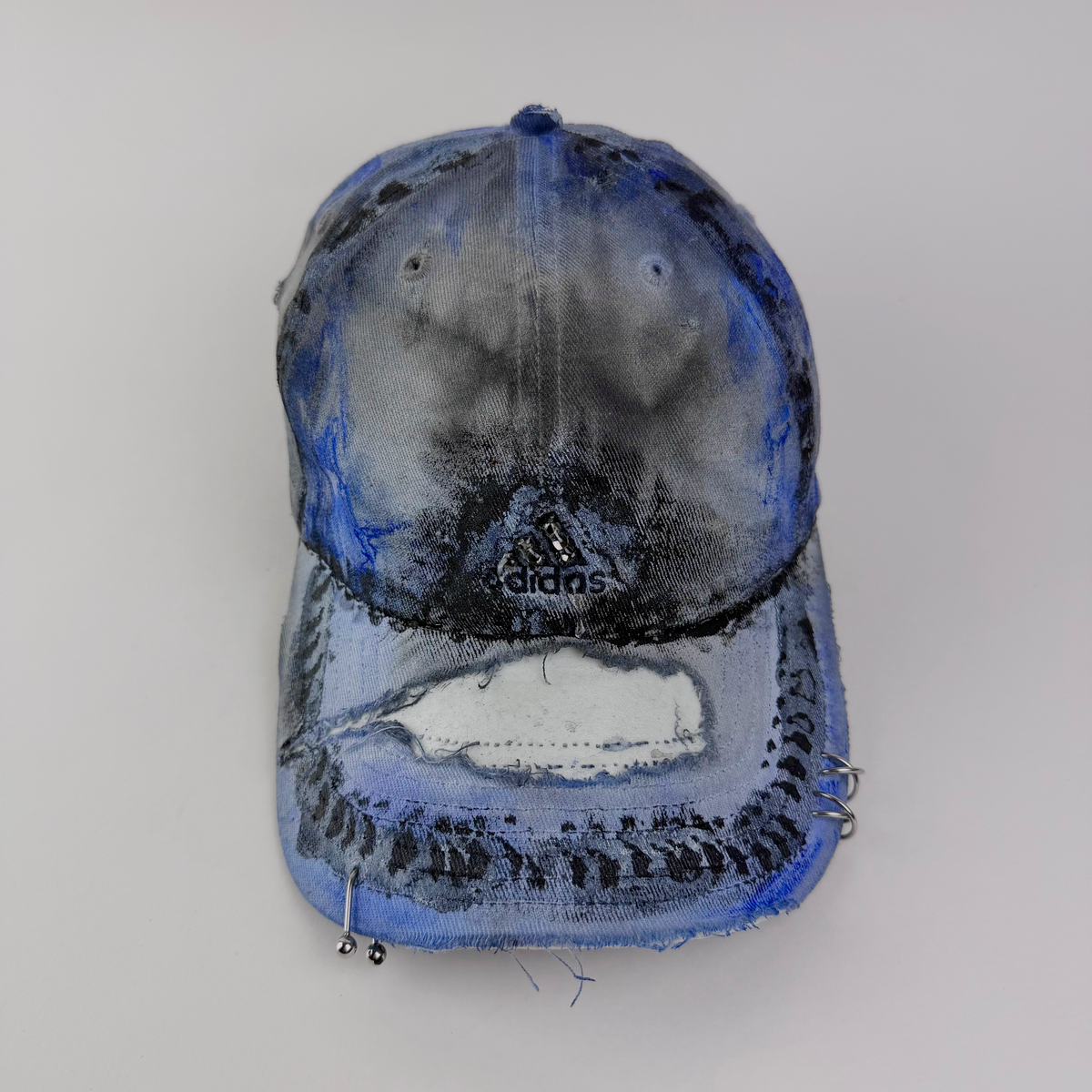 ASTROBOI* x Adidas 'Psykos' white blue and black tie-dye painted grunge cap with rhinestone-embellished Adidas logo, black and blue faded abstract design, stainless steel piercings, and deconstructed visor, front view.