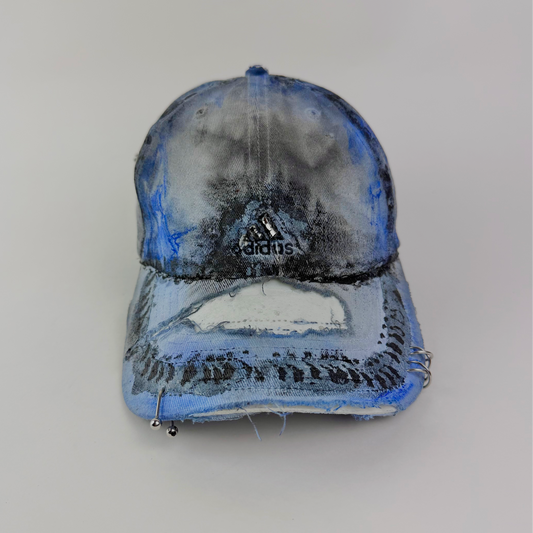ASTROBOI* x Adidas 'Psykos' white blue and black tie-dye painted grunge cap with rhinestone-embellished Adidas logo, black and blue faded abstract design, stainless steel piercings, and deconstructed visor, front view.