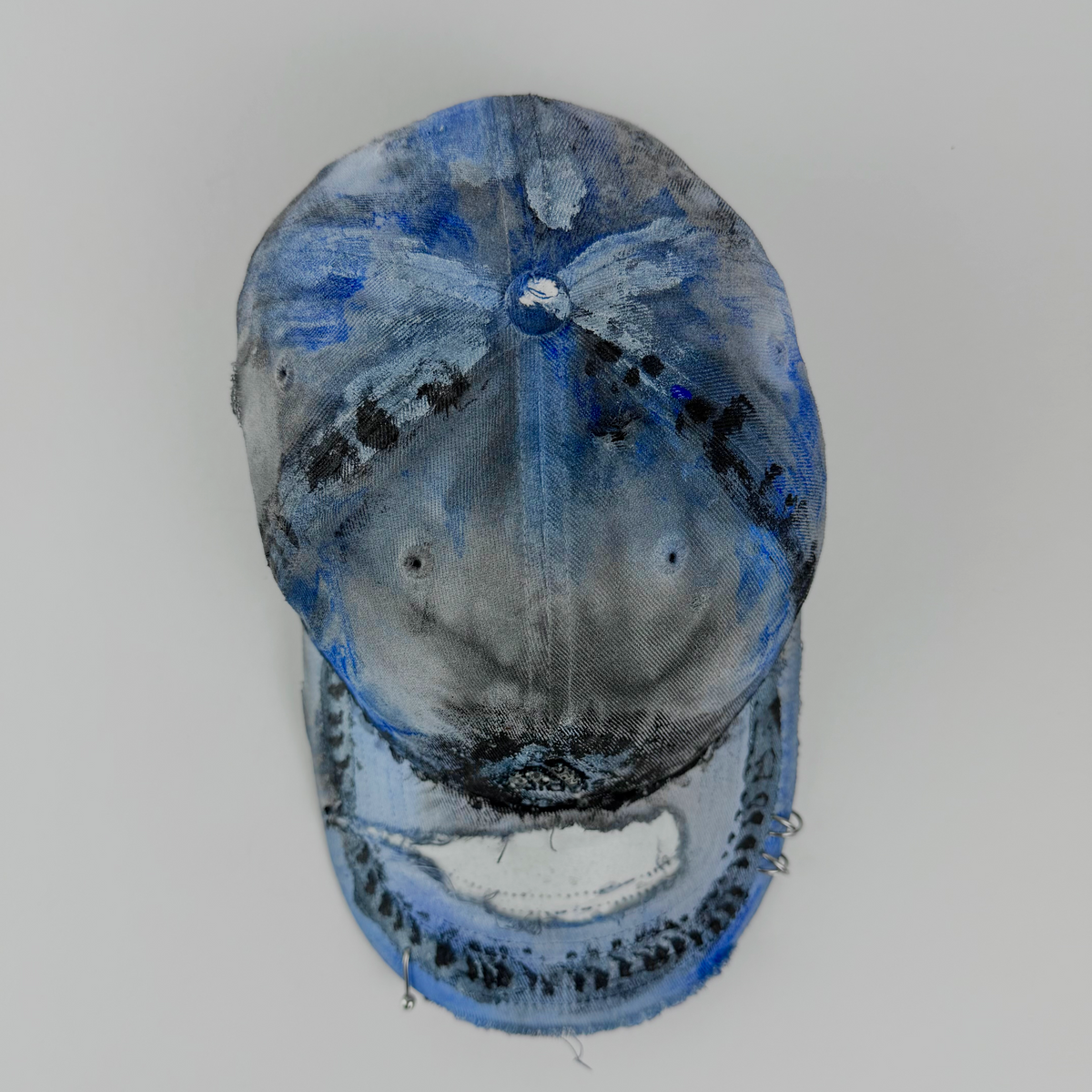 A distressed baseball cap with a heavily worn, painted finish in shades of blue, gray, and black, photographed from a top-down angle against a light gray background.