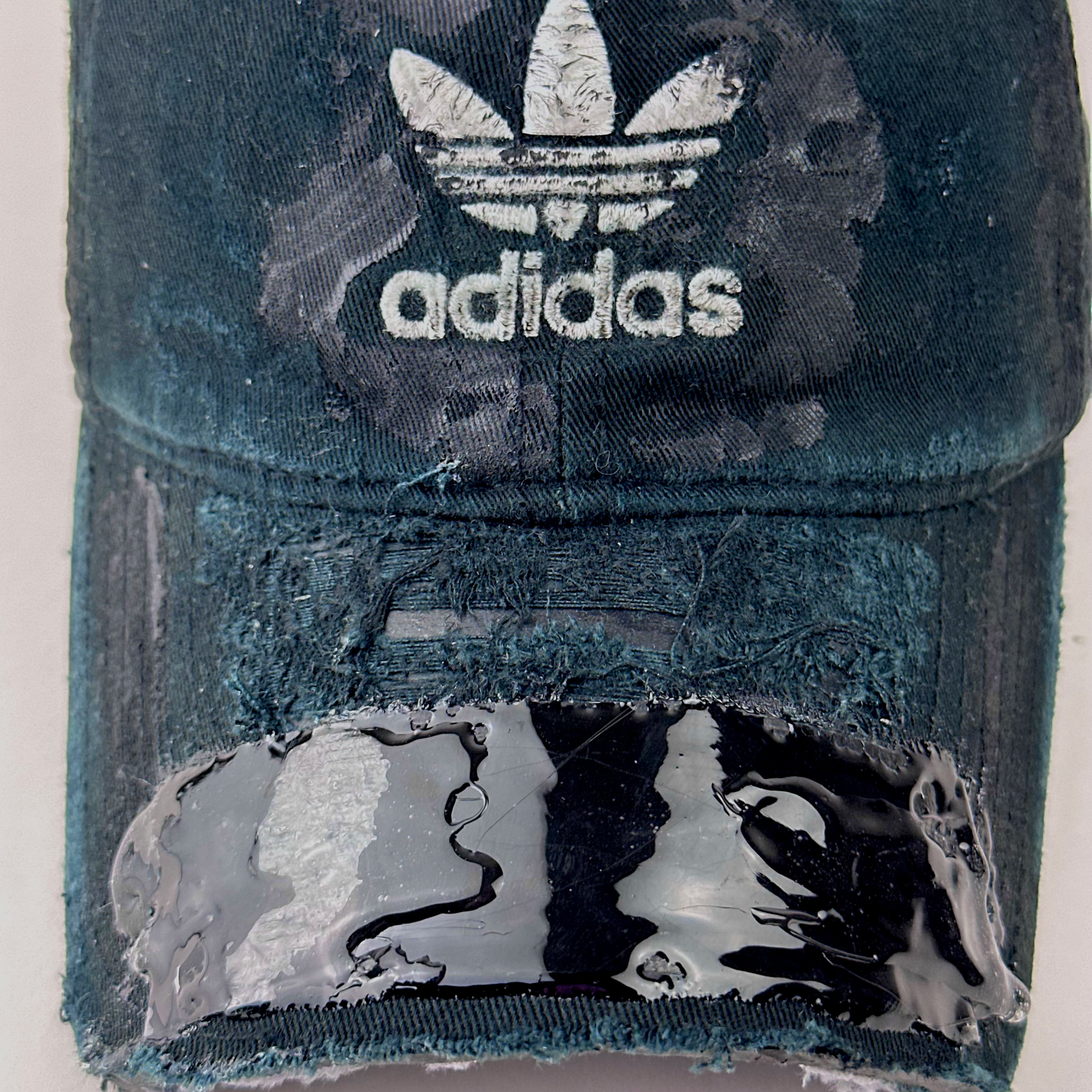 Close-up of ASTROBOI* x Adidas 'Washed' showing Adidas trefoil logo, black to navy fade, and melted duct tape sealed under resin on partially sawed-off visor.