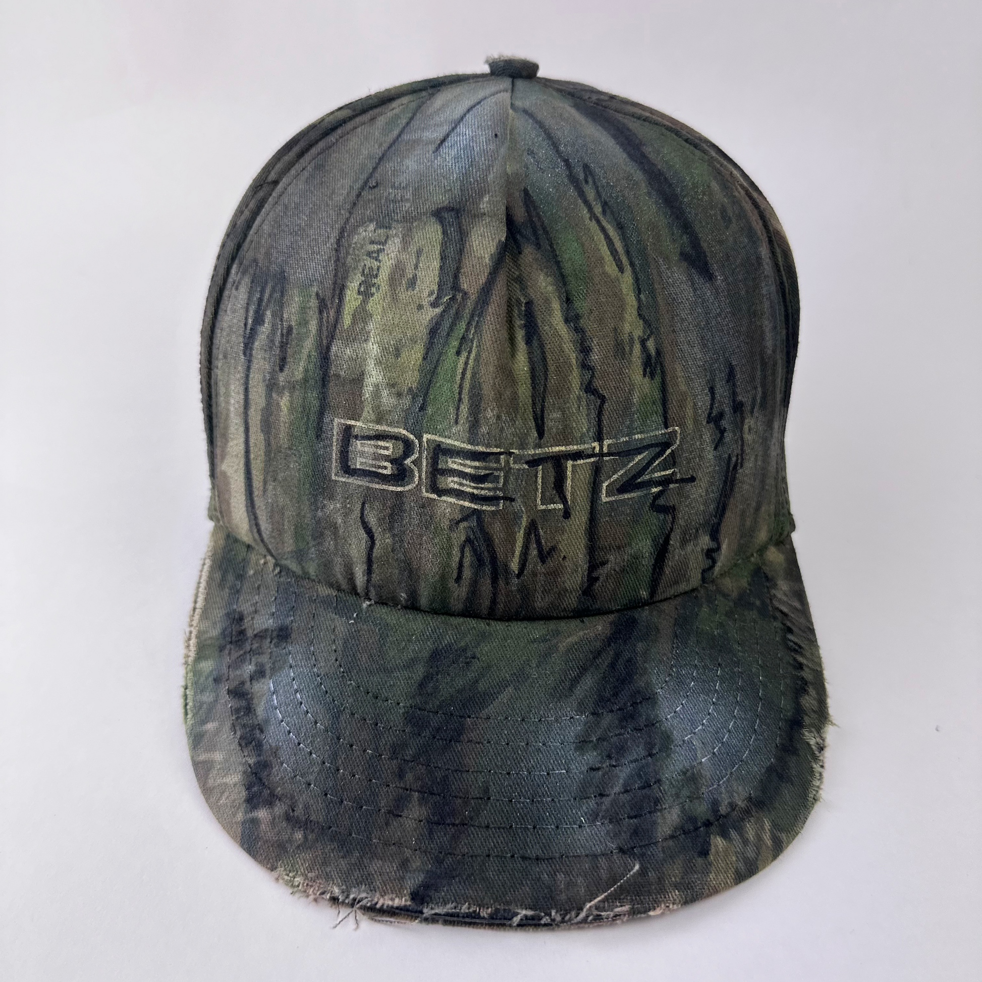 ASTROBOI* x Betz brown Real Tree camouflage doodle hat with hand drawn design, custom paint treatment, and Betz logo at front, front view.