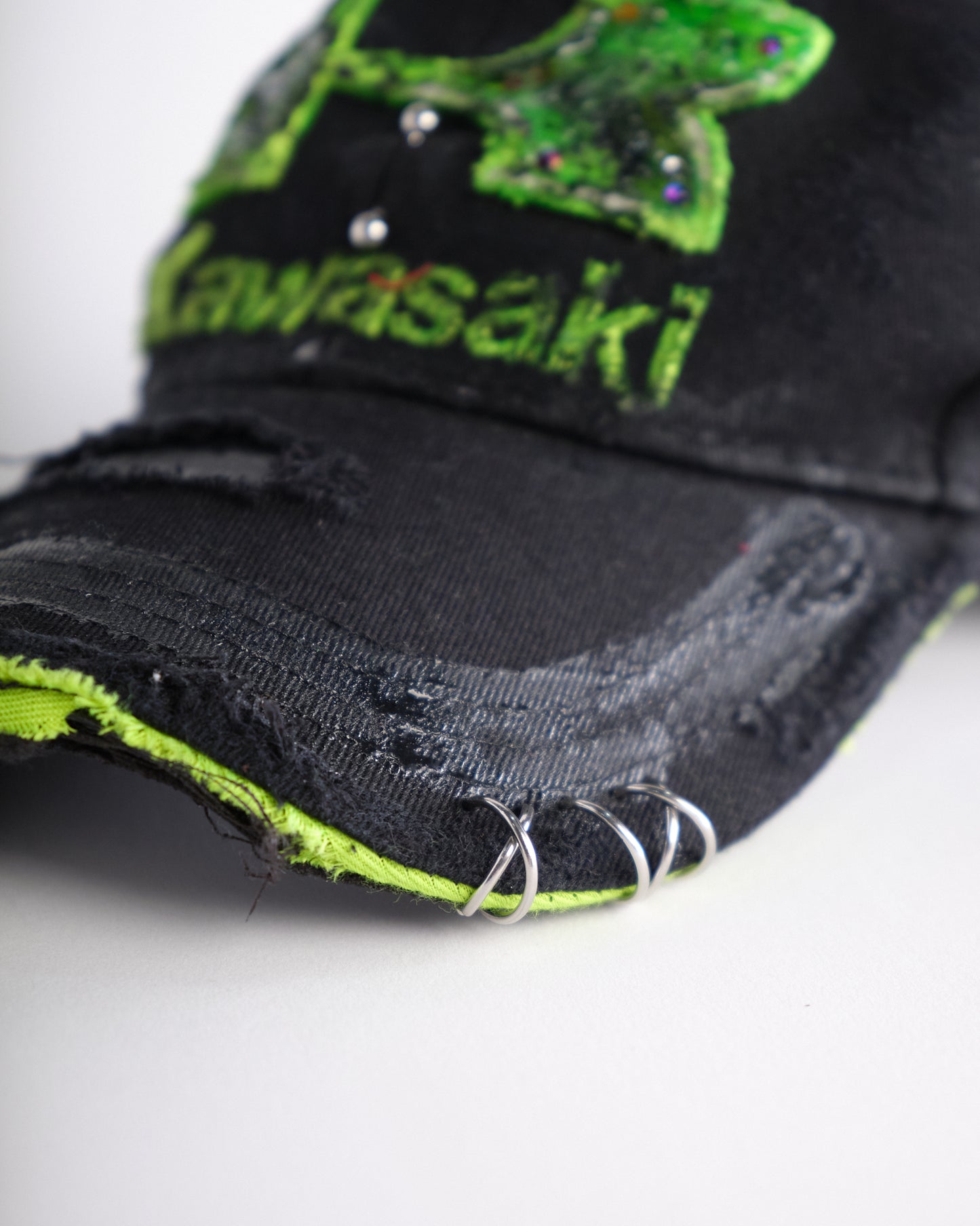 Piercing details on ASTROBOI* x Kawasaki black hat with neon green rhinestone logo, resin finish, black paint distressing, and stainless steel piercings.