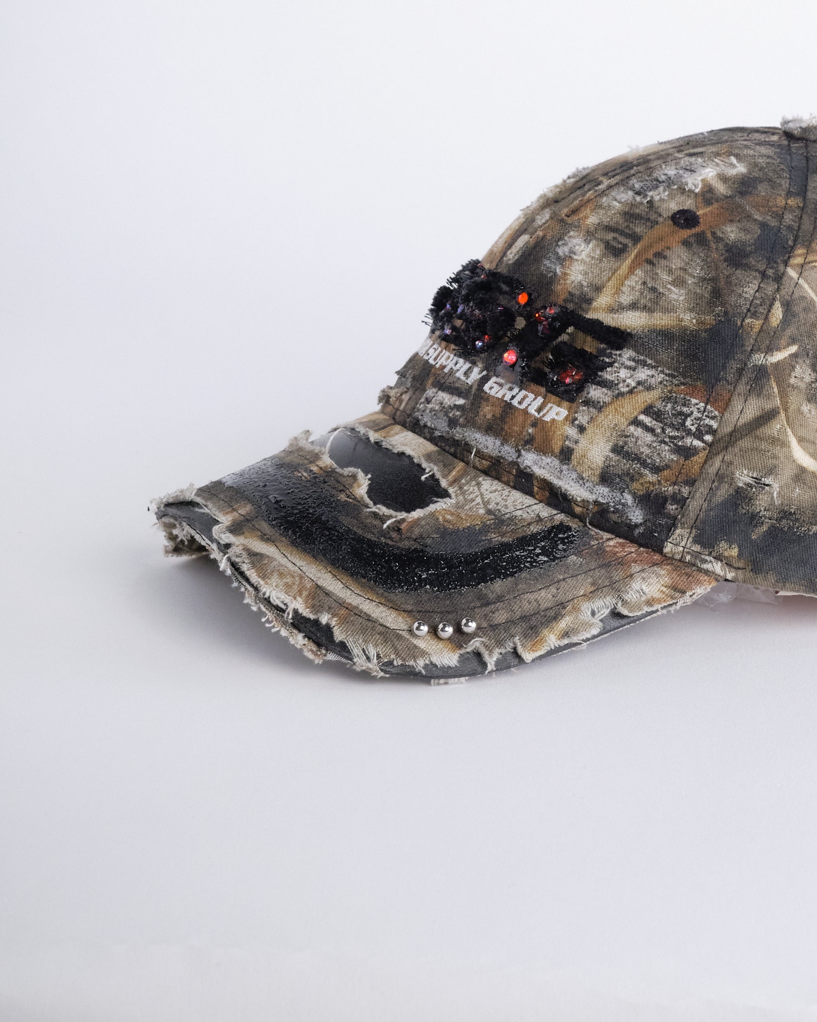 Front visor detail of ASTROBOI* DSG brown camouflage hat with rhinestone logo, black paint, clear resin accents, and stainless steel piercings, adjustable fit, handmade in NYC.