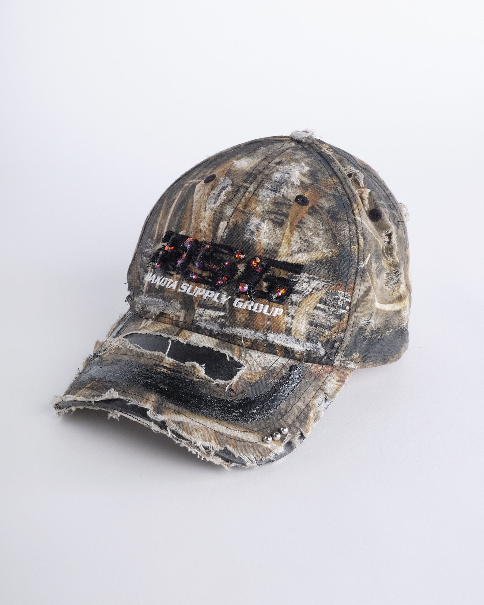 Front left side of ASTROBOI* DSG brown camouflage hat with rhinestone logo, black paint, clear resin accents, and stainless steel piercings, adjustable fit, handmade in NYC.