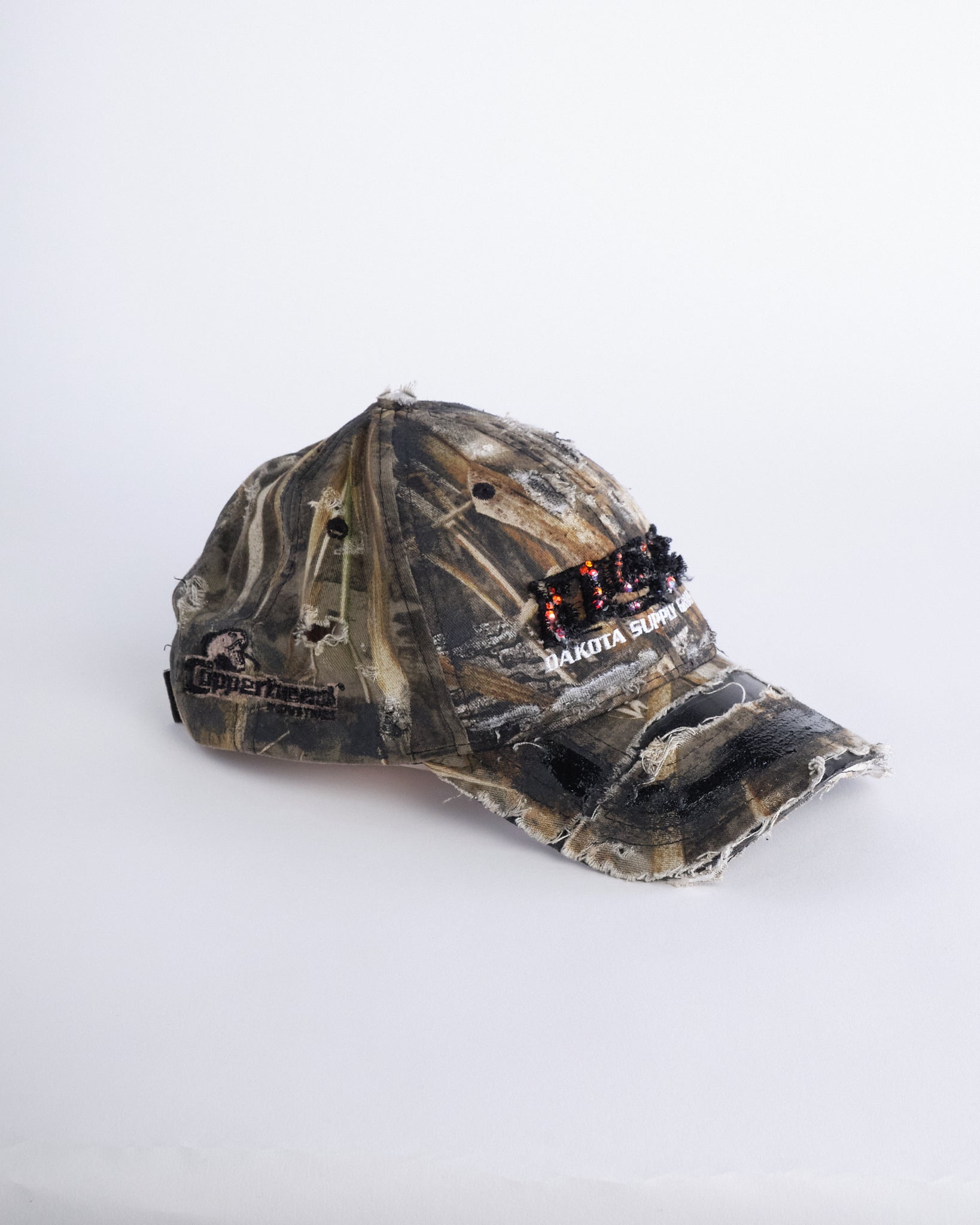 Right side of ASTROBOI* DSG brown camouflage hat with rhinestone logo, black paint, clear resin accents, and stainless steel piercings, adjustable fit, handmade in NYC.