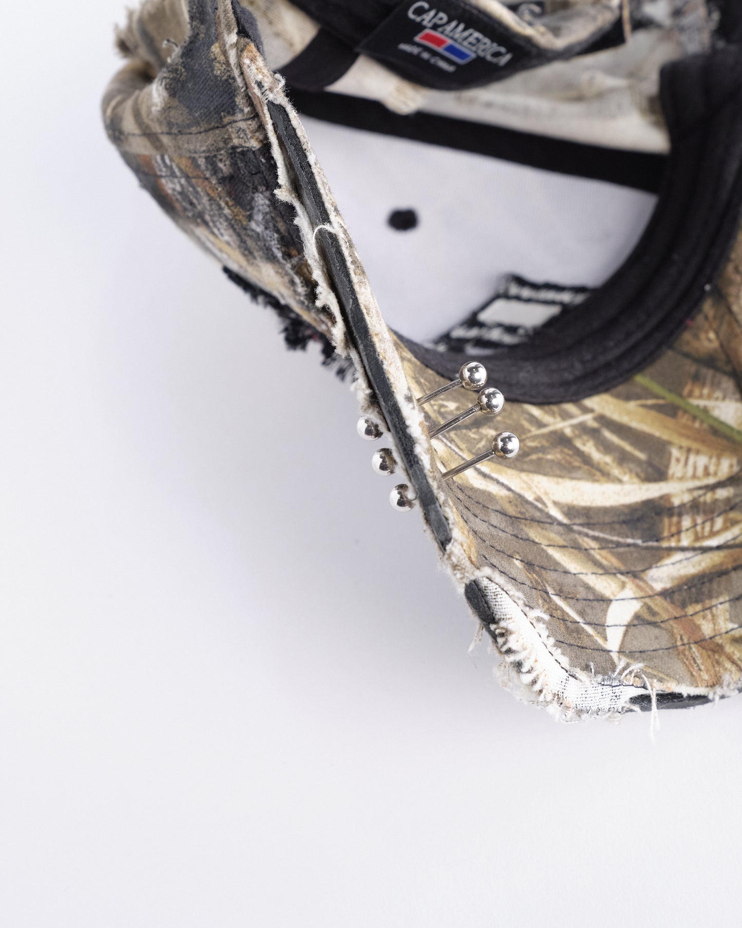 Visor detail of ASTROBOI* DSG brown camouflage hat with rhinestone logo, black paint, clear resin accents, and stainless steel piercings, adjustable fit, handmade in NYC.