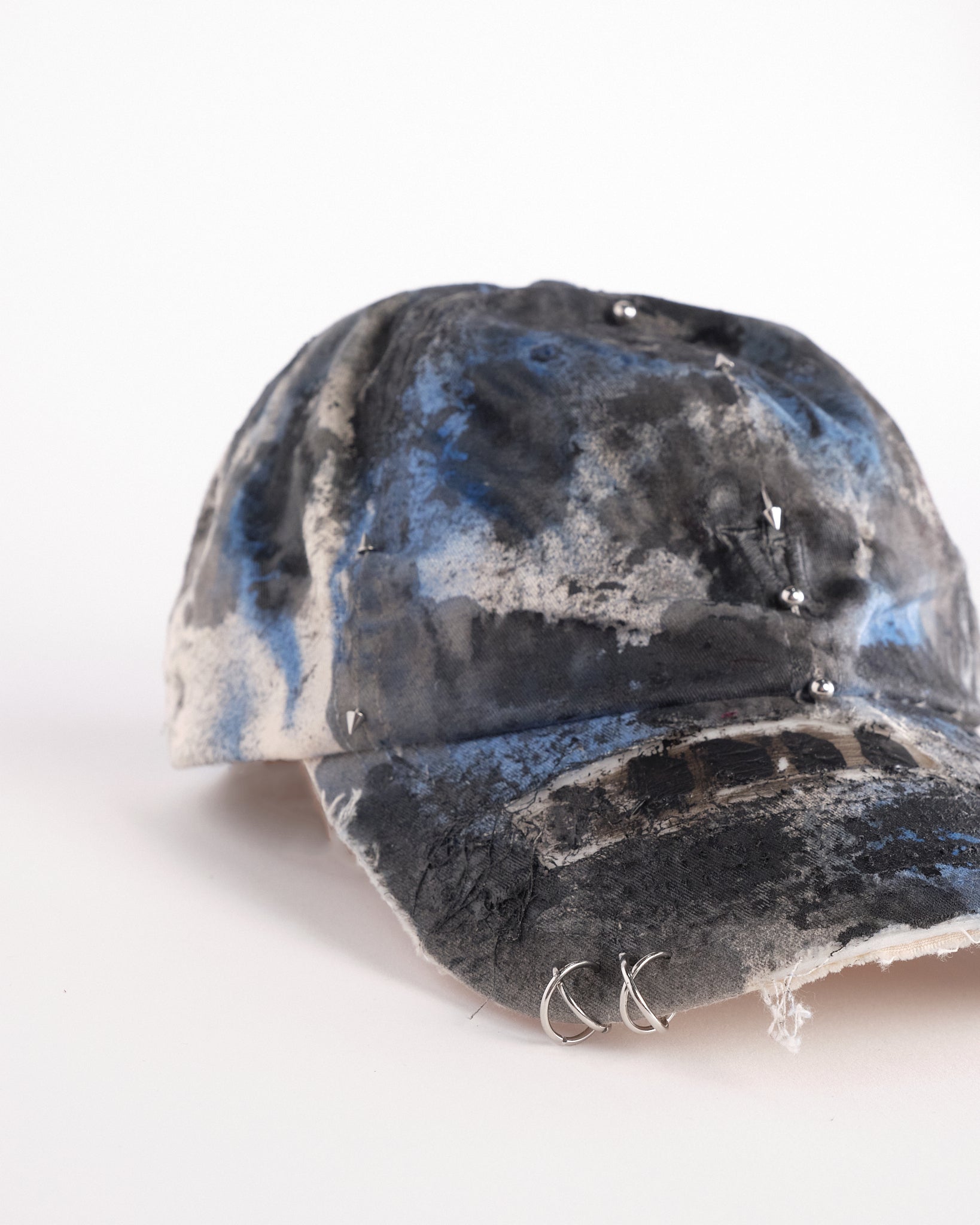 Front visor detail of Black Vuori baseball hat customized by ASTROBOI* with blue and black painted details, stainless steel piercings, and an embroidered V logo.