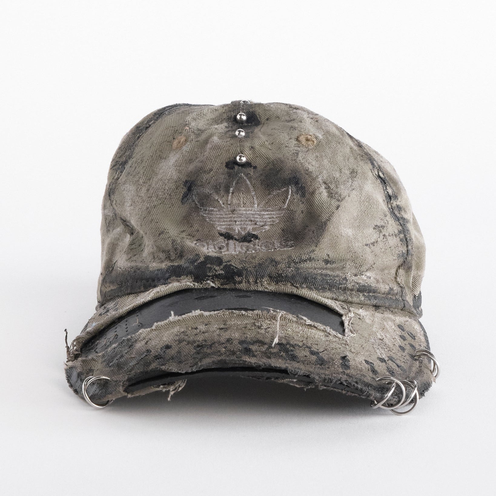 Front of ASTROBOI* x Adidas tan distressed hat with black paint detailing, Adidas logo at front, and stainless steel piercings, customized in NYC.