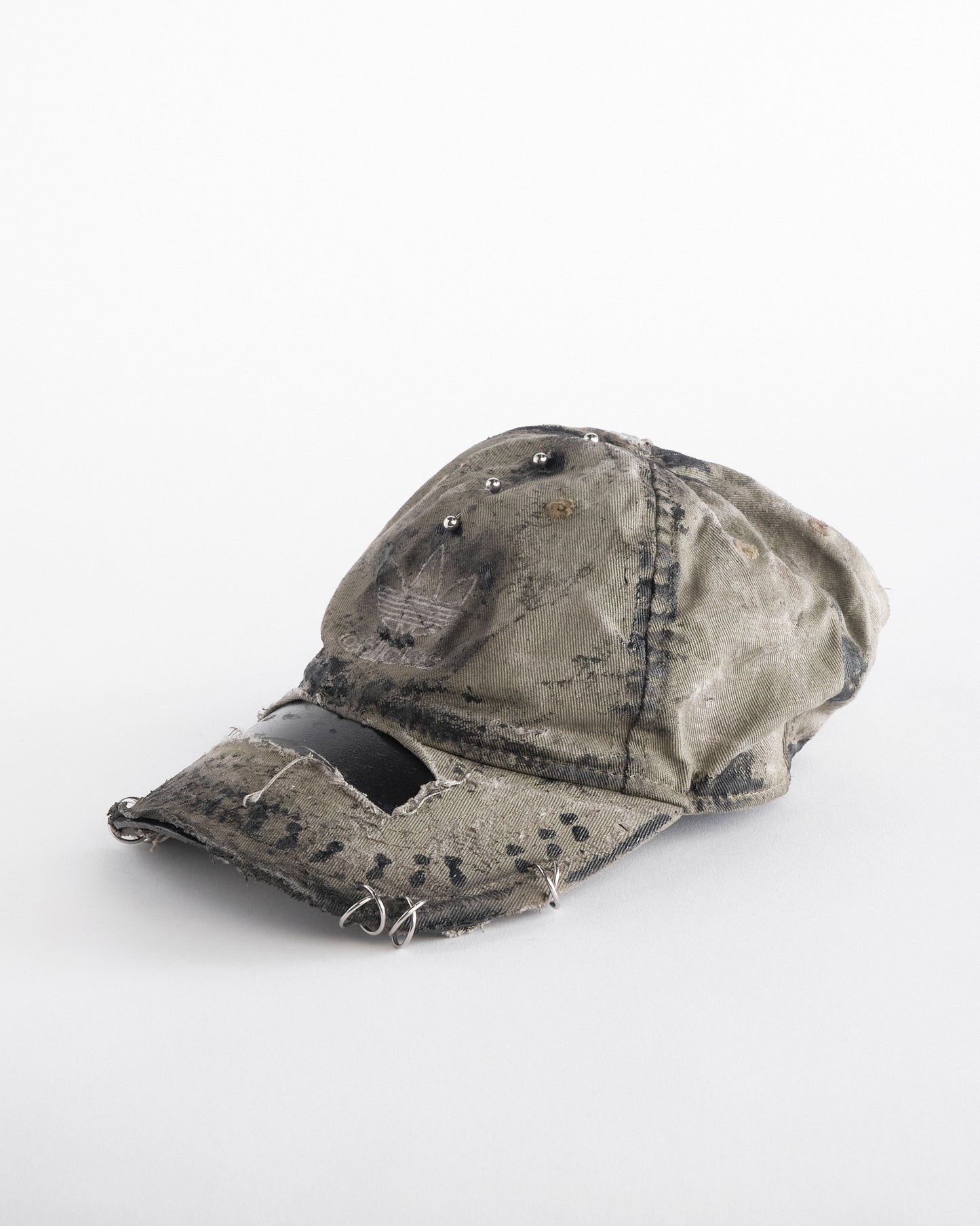 Front left of ASTROBOI* x Adidas tan distressed hat with black paint detailing, Adidas logo at front, and stainless steel piercings, customized in NYC.