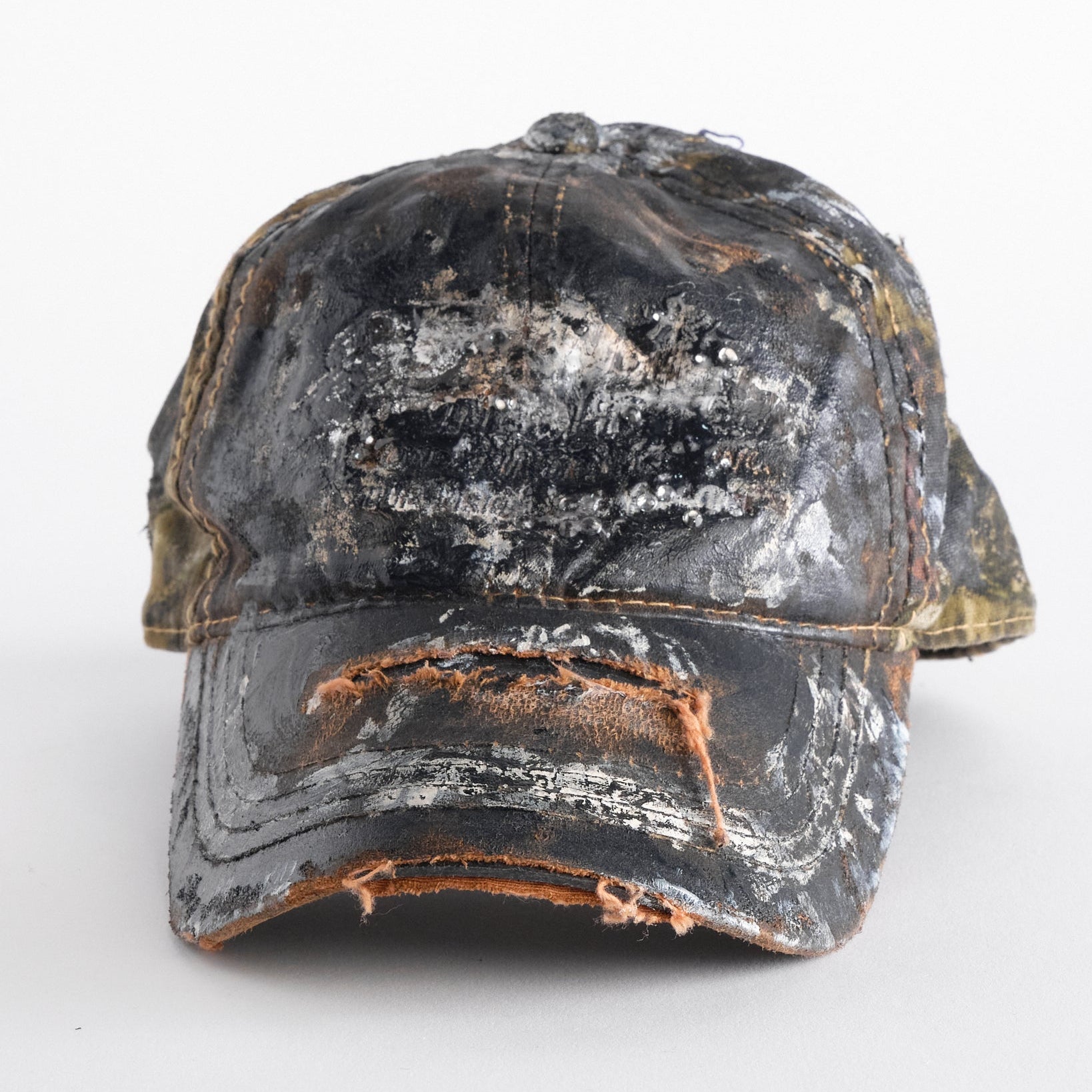 Front of ASTROBOI* x Mossy Oak brown RealTree camo hat with rhinestones, distressed paint details, and leather visor, adjustable fit.