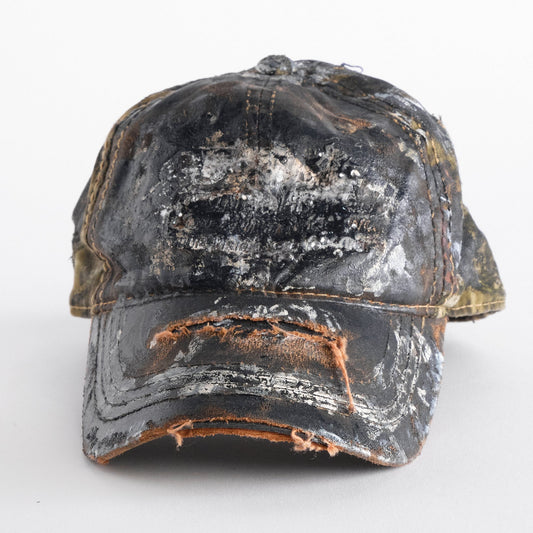 Front of ASTROBOI* x Mossy Oak brown RealTree camo hat with rhinestones, distressed paint details, and leather visor, adjustable fit.