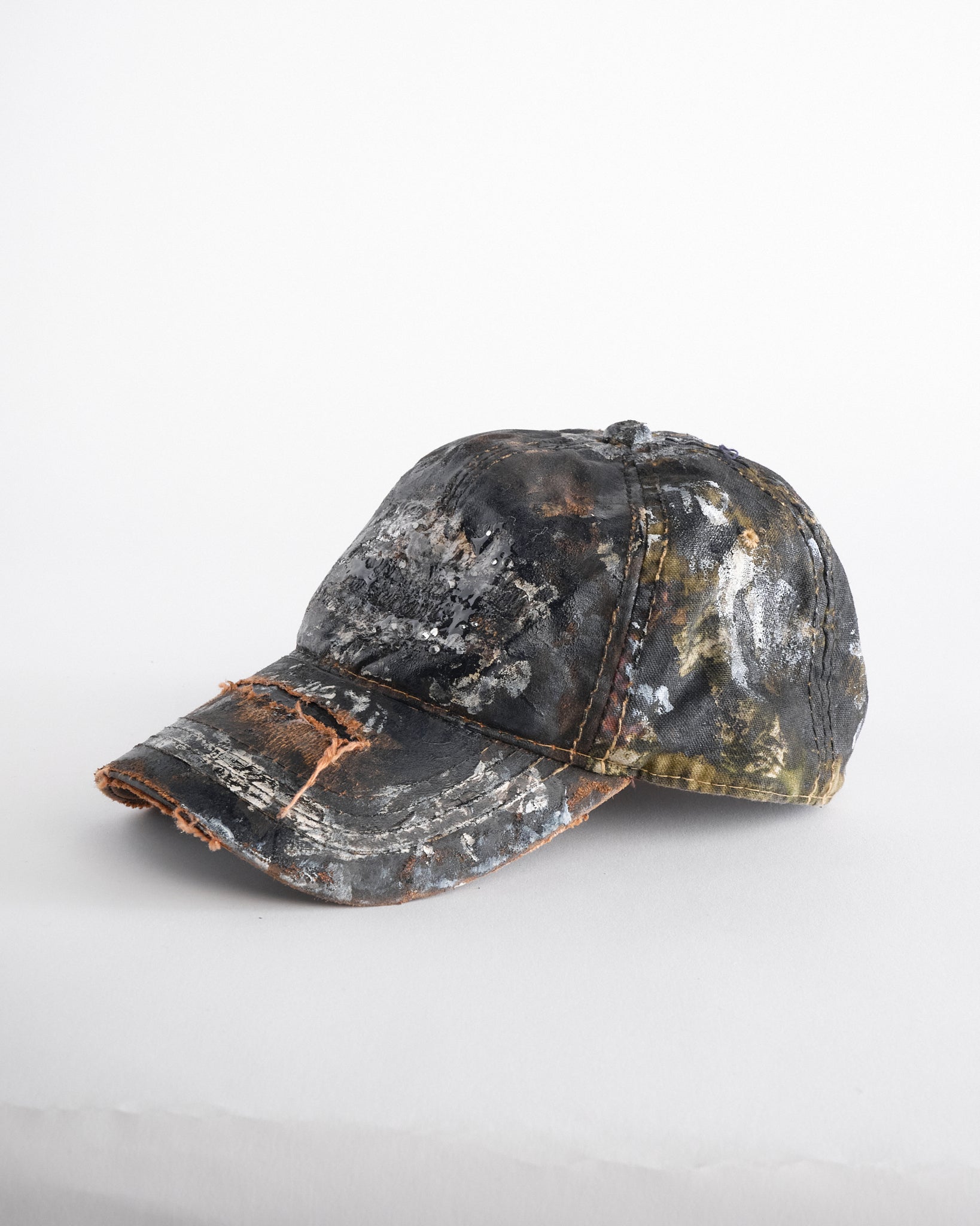 Left of ASTROBOI* x Mossy Oak brown RealTree camo hat with rhinestones, distressed paint details, and leather visor, adjustable fit.