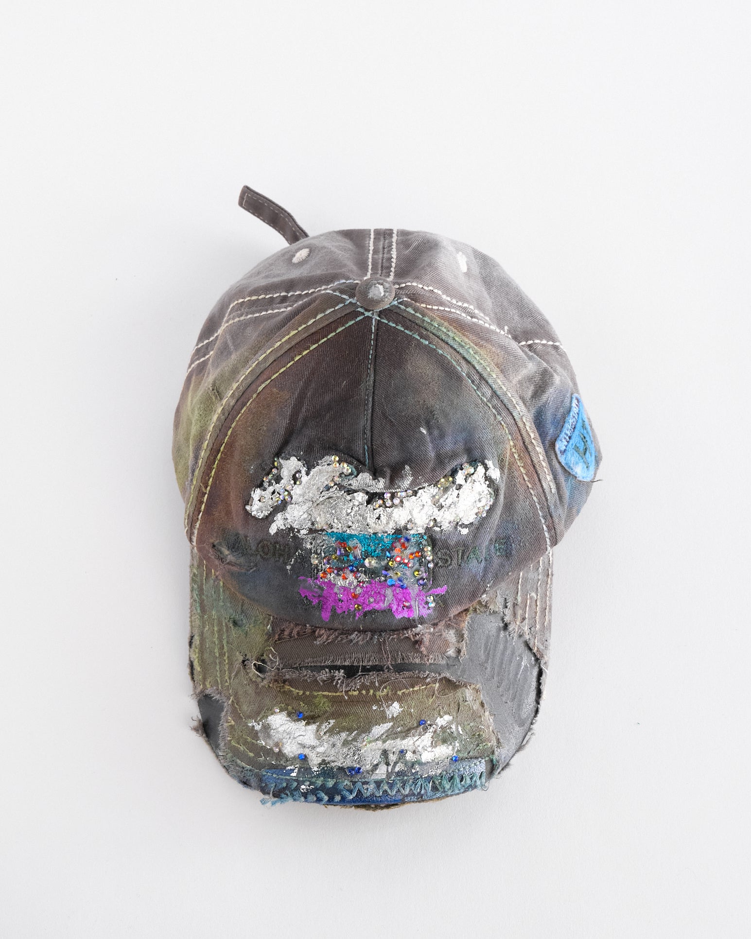 Overview of Brown-green ASTROBOI* hat featuring a metallic Hawaii logo with multicolored rhinestones, distressed visor, and resin-coated safety pin detail.