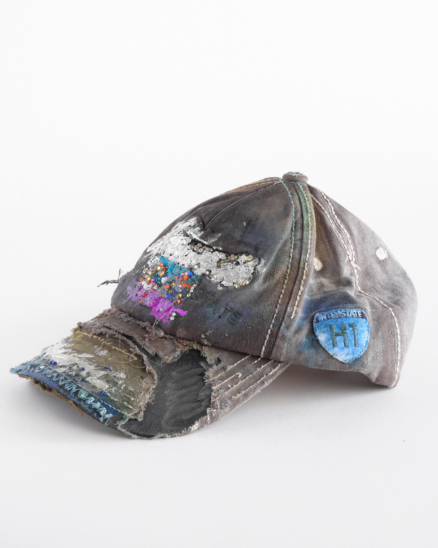 Left of Brown-green ASTROBOI* hat featuring a metallic Hawaii logo with multicolored rhinestones, distressed visor, and resin-coated safety pin detail.