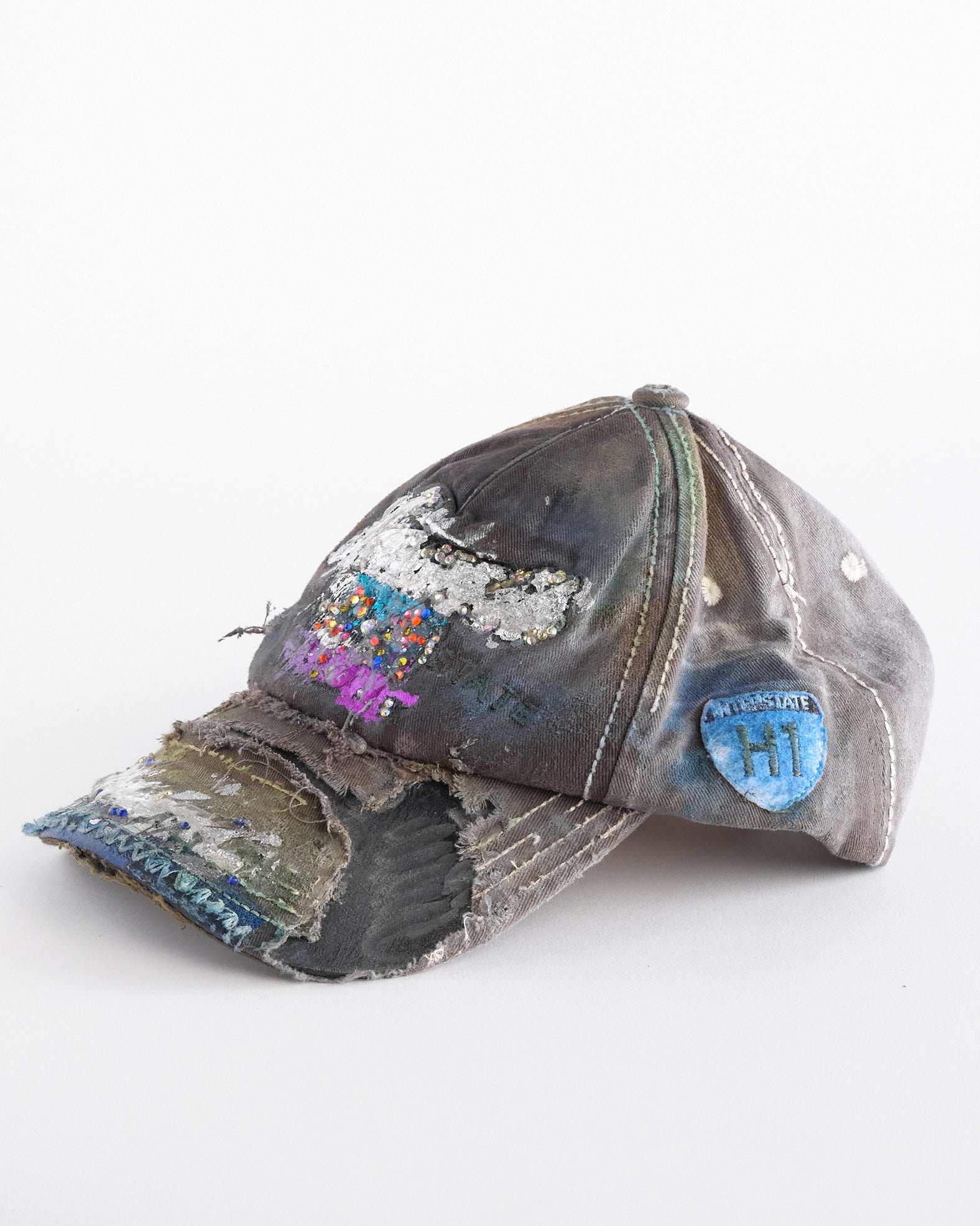 Left of Brown-green ASTROBOI* hat featuring a metallic Hawaii logo with multicolored rhinestones, distressed visor, and resin-coated safety pin detail.