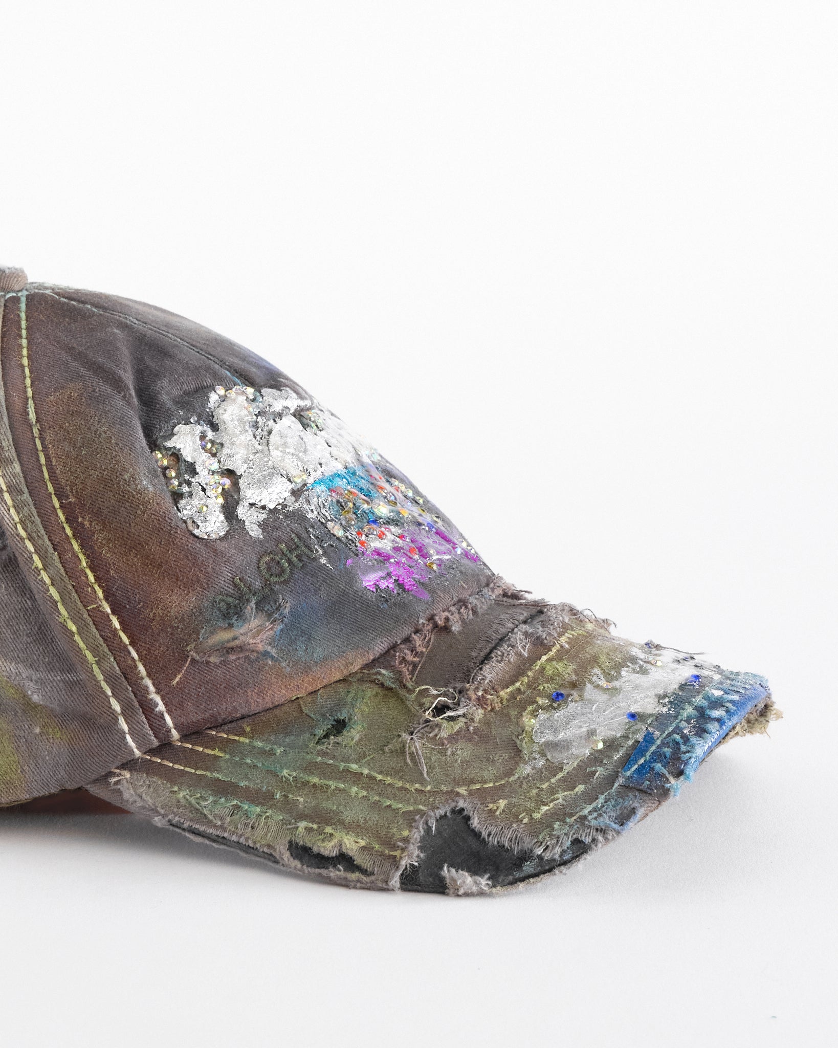 Visor detail of Brown-green ASTROBOI* hat featuring a metallic Hawaii logo with multicolored rhinestones, distressed visor, and resin-coated safety pin detail.