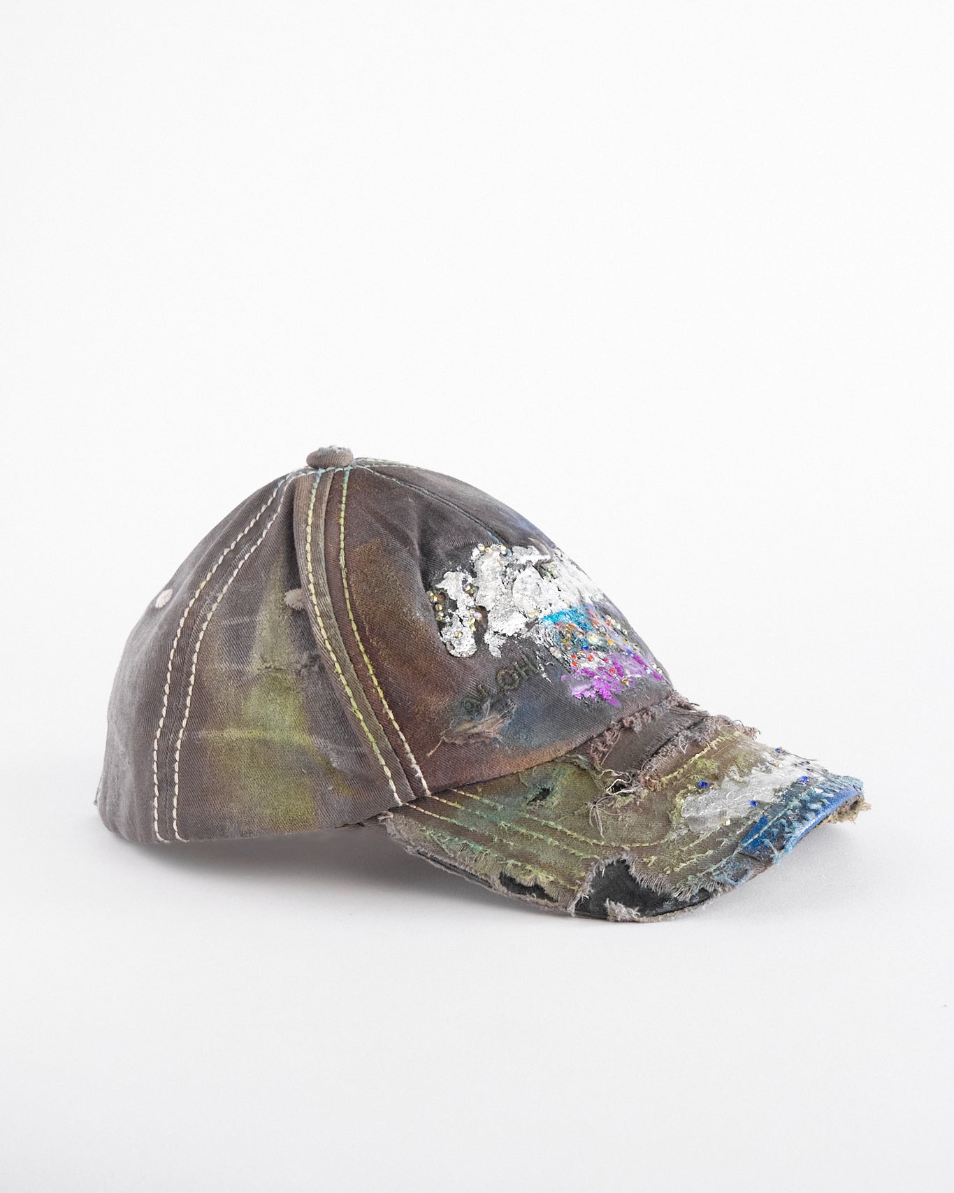 Right of Brown-green ASTROBOI* hat featuring a metallic Hawaii logo with multicolored rhinestones, distressed visor, and resin-coated safety pin detail.