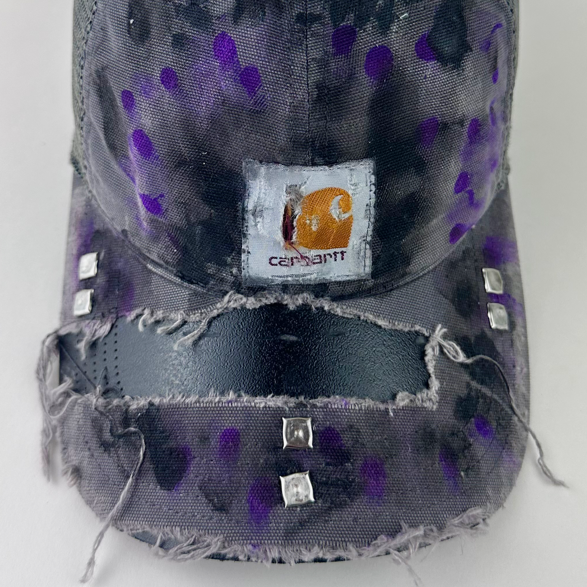 Close-up of ASTROBOI* x Carhartt 'Amethyst' showing Carhartt logo patch and silver stud detail on deconstructed visor with black and purple paint treatment.