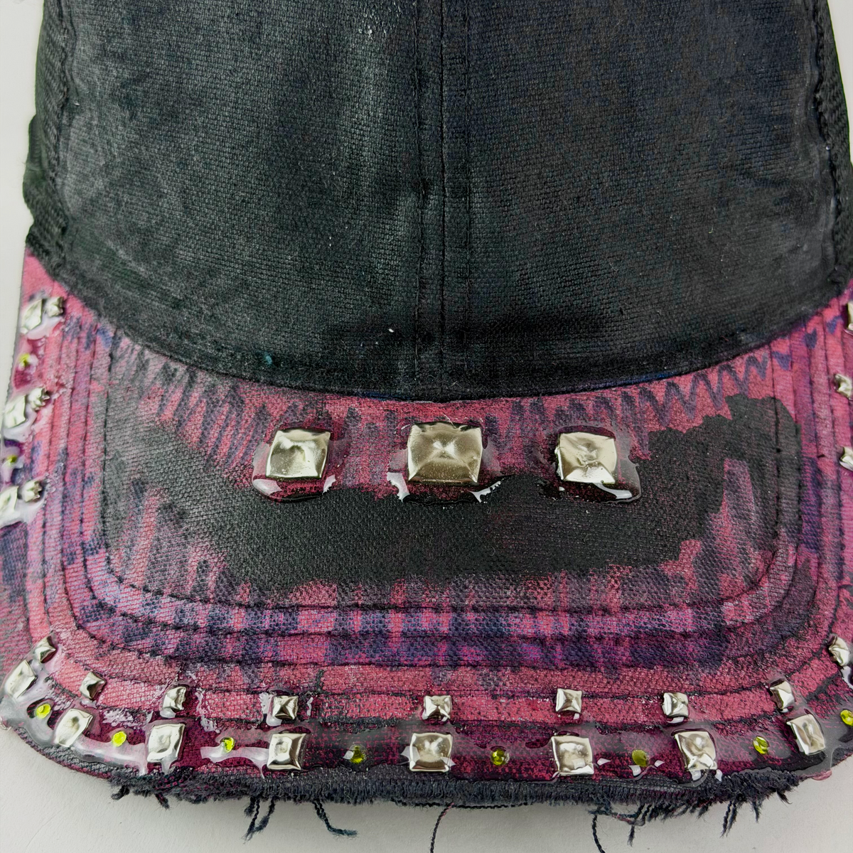 A close-up of a distressed baseball cap with a black crown and a brim featuring a pink and purple tie-dye pattern, heavily embellished with silver metal studs and rhinestones.