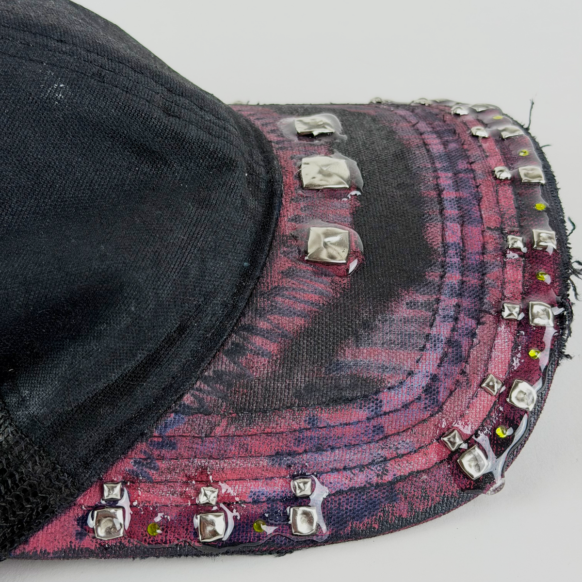 A close-up of a baseball cap featuring a black fabric crown and a curved brim decorated with a distressed pink and purple pattern, embellished with multiple silver-tone metal studs and rhinestones.
