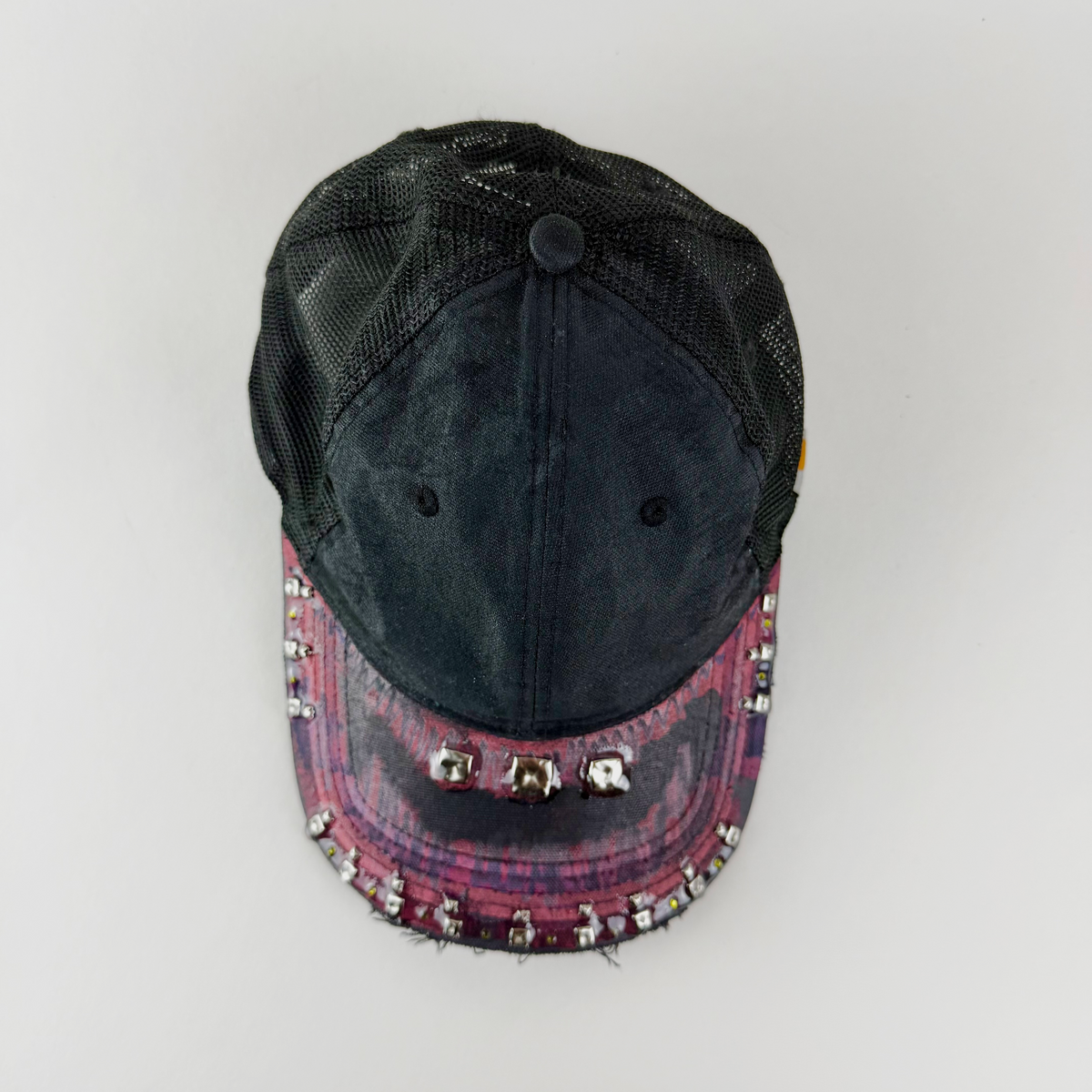 A distressed trucker-style baseball cap with a black mesh back and a frayed, patterned brim in pink, purple, and black, embellished with rhinestones and silver studs.