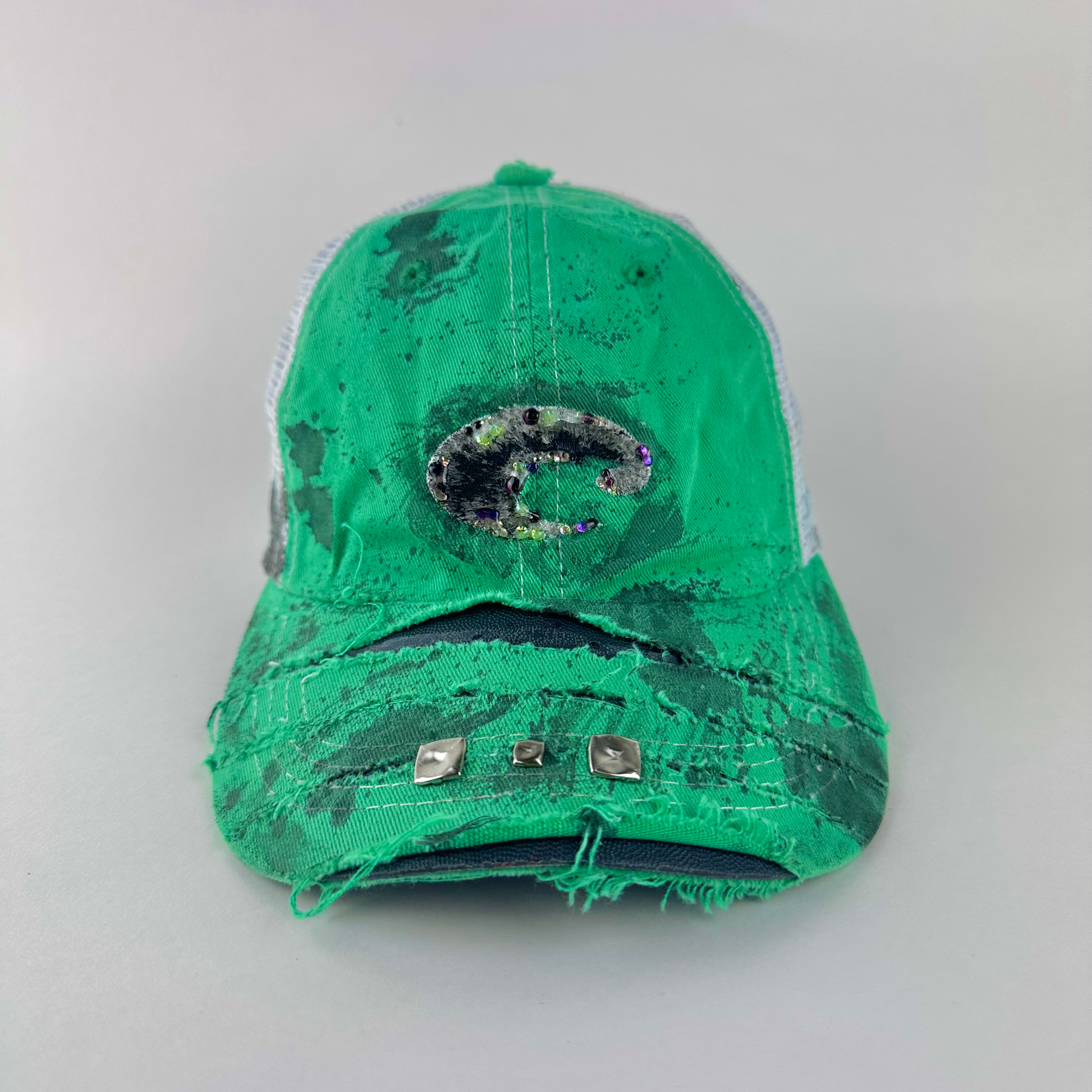 ASTROBOI* x Costa del Mar 'Jade' Y2K green rhinestone studded trucker hat with purple and green rhinestone-embellished Costa 'C' logo, silver studs on deconstructed visor, and black stained paint treatment, front view.