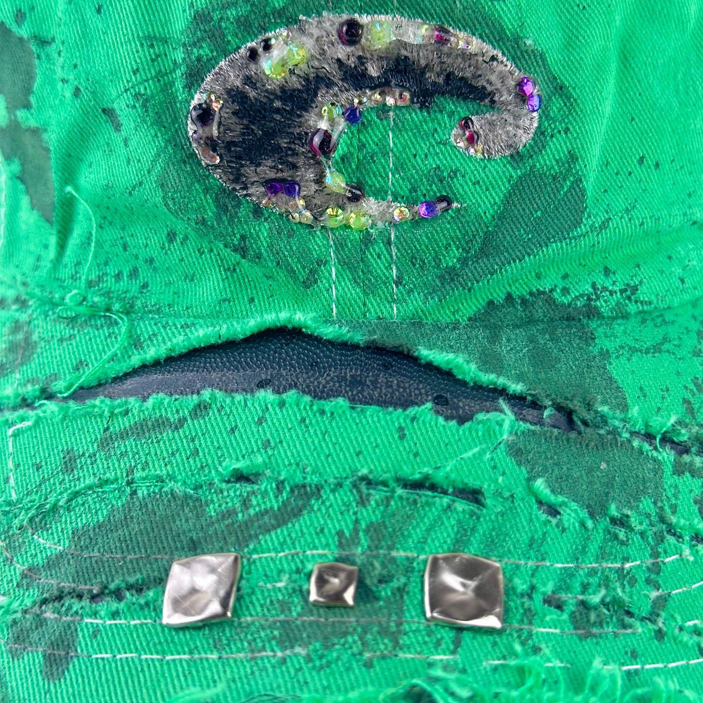 Close-up of a bright green distressed denim fabric with a large metallic silver and black patch, frayed edges, and three silver metal studs on a white background.