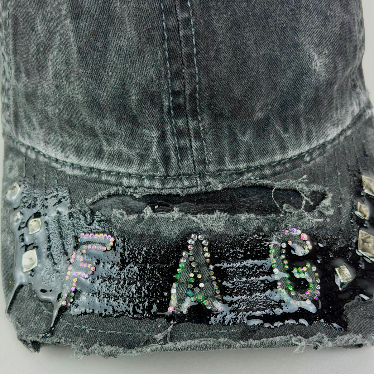 A close-up of a distressed black denim baseball cap with 'FAG' spelled out in colorful rhinestone letters on the brim, set against a plain light gray background.