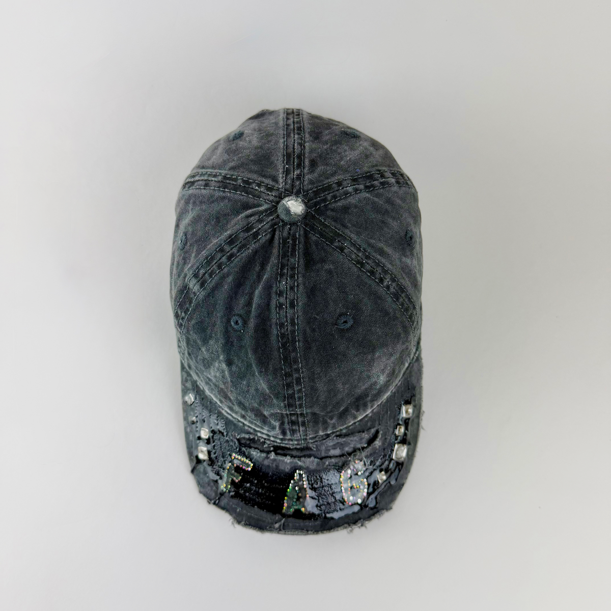 A washed black denim baseball cap with rhinestone embellishments spelling 'FAG' on the brim, photographed from a top-down angle against a light gray background.